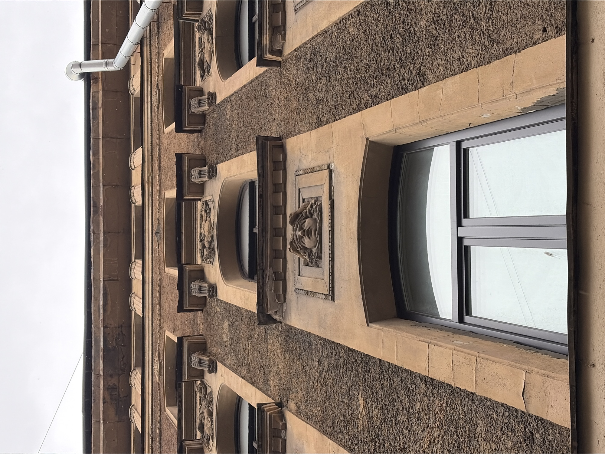 The image shows a close-up, low-angle view of the upper facade of a building in Saint Petersburg, Russia, likely an older apartment building or historic structure. The facade is predominantly constructed of textured, light brown stucco with lighter, smooth stone lintels and decorative elements.
Key architectural features visible include:
* **Windows:** Several arched windows with dark gray frames are prominent. The glass reflects the overcast sky and faint outlines of buildings or objects outside.
* **Decorative Reliefs:**
* A central, prominent relief of a human face, possibly a mask or a bust, is located above a larger window. The face is detailed with curly hair and a serene expression. It is framed by a rectangular relief with a decorative border.
* Smaller decorative reliefs, appearing to be figures or scenes, are situated above some of the upper arched windows.
* Ornate corbels or brackets are present, supporting architectural projections above some of the windows.
* **Cornices and Projections:** A distinct, layered cornice runs along the top of the building, with a flat, dark roofline visible above it.
* **Downspout:** A silver metal downspout extends diagonally from the upper right of the frame, attached to the building's facade.
* **Texture:** The contrast between the rough, pebbled texture of the main stucco and the smooth stone elements is a defining characteristic.
The overcast sky suggests it is a daytime shot. The perspective emphasizes the verticality and the ornate details of the building's upper story. The building appears to be located in an urban environment, typical of Saint Petersburg's historic districts. The overall impression is one of architectural grandeur and historical significance, with a slightly weathered patina.