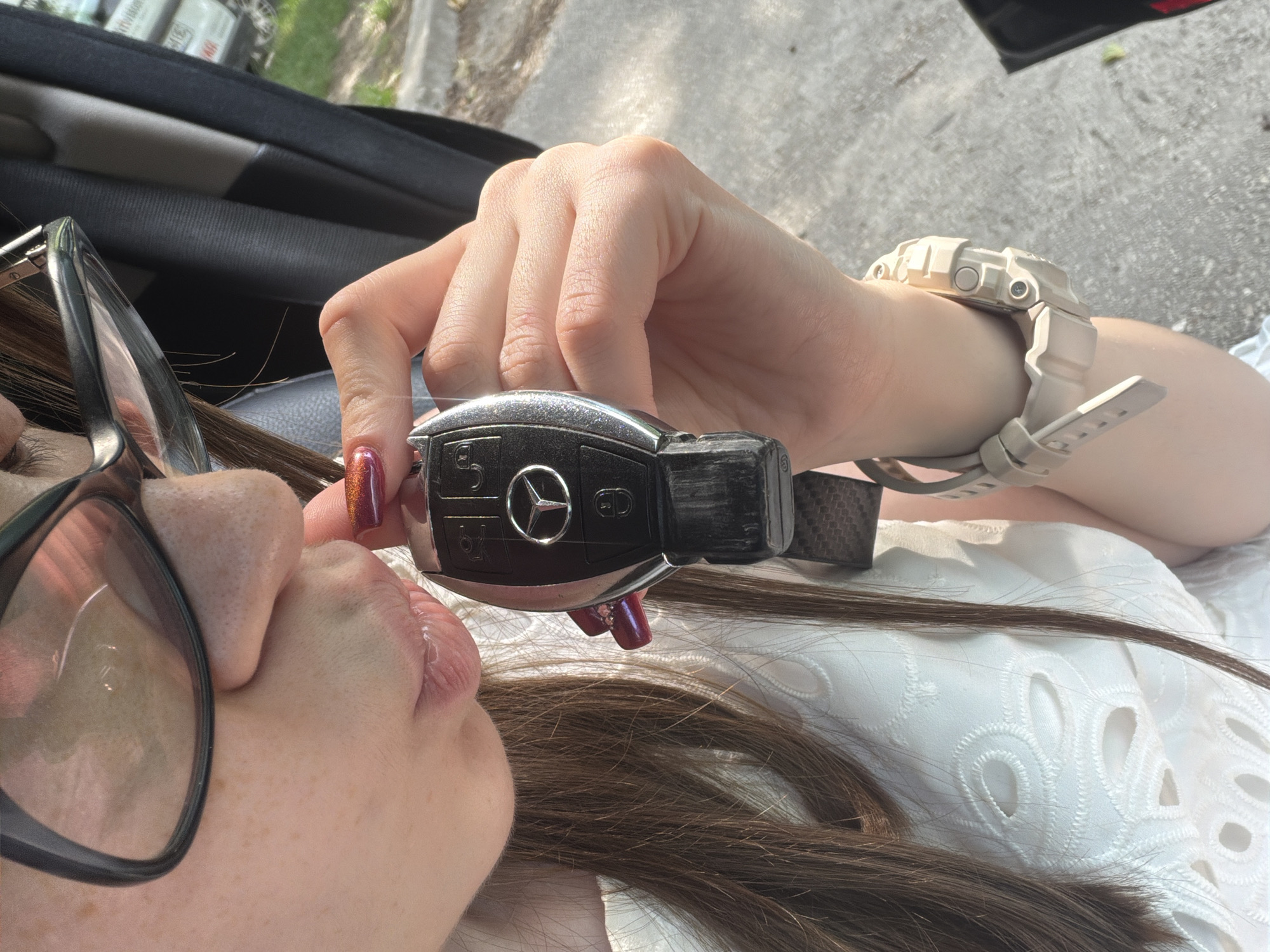 The image presents a close-up, eye-level view of a female individual interacting with a car key. The subject, observed from the nose down, has long brown hair cascading over a white garment featuring an embroidered eyelet pattern. Dark-framed eyeglasses are worn, exhibiting a reflection on the right lens. The subject's lips, slightly parted and glossy, are positioned directly in front of a black Mercedes-Benz car key. This key is held by the subject's right hand; the fingernails are adorned with a metallic orange-red polish, and the index finger shows a small, dark decorative element near the cuticle. On the subject's left wrist, a light beige/sand-colored G-Shock digital wristwatch is visible. The immediate background indicates a vehicle interior, including a dark seatbelt strap. Beyond this, an outdoor environment is discernible, featuring an asphalt surface, minimal green vegetation, and distant parked vehicles under bright daylight. A white vehicle in the far background displays partially visible text, specifically "Varna" or "Варна", confirming the scene's location within Varna, Bulgaria.