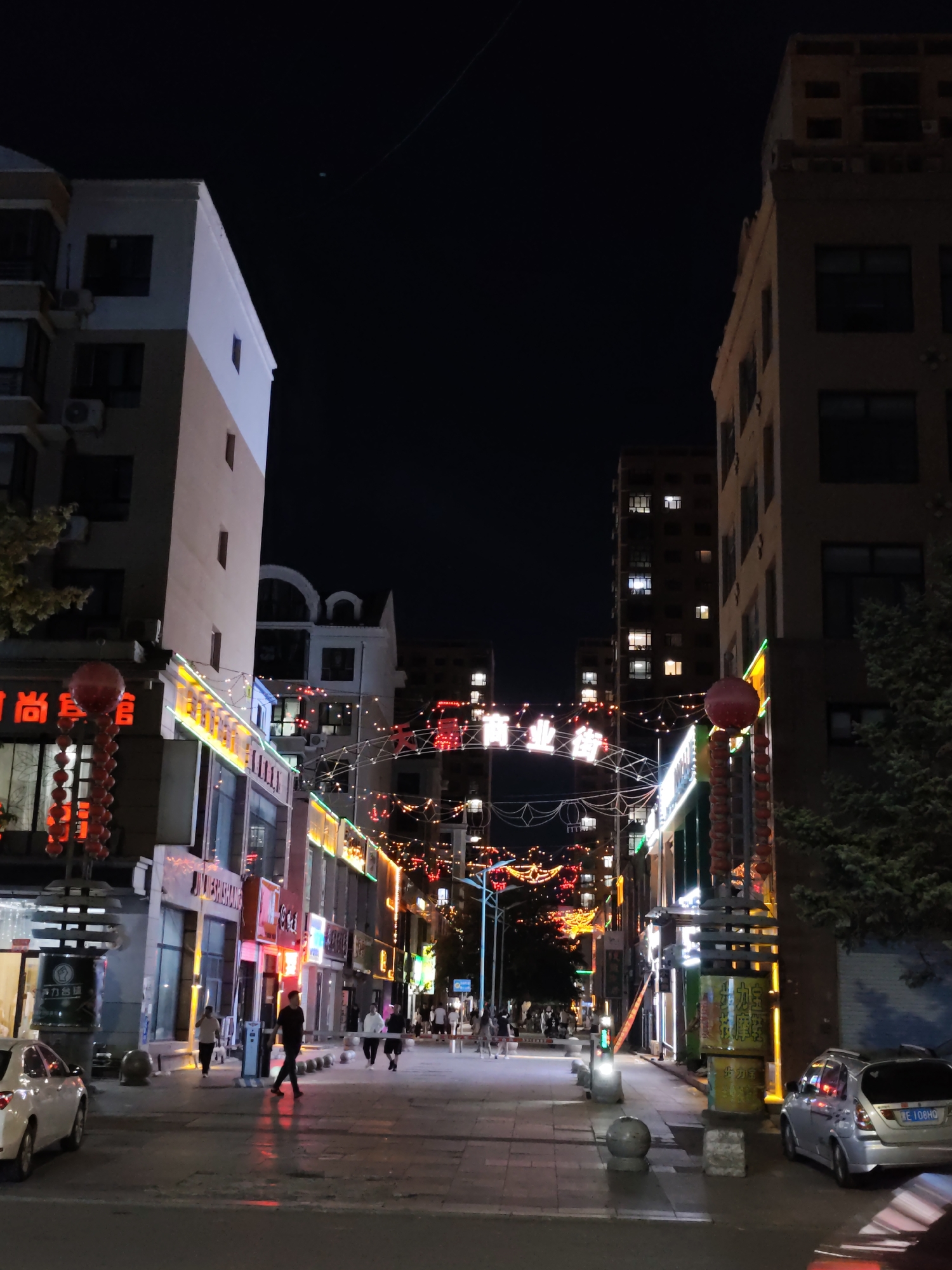 The image depicts a street scene at night in Lindian, China, with buildings lining both sides of the thoroughfare. The street is illuminated by a variety of lights, including neon signs, string lights, and traffic lights, creating a festive atmosphere. Several buildings appear to be commercial establishments, indicated by the illuminated signs. Some people are walking on the sidewalks and crossing the street. Two cars are parked on the side of the road. Red lanterns and decorative lights are suspended across the street, adding to the ambiance. The sky is dark, indicating it is nighttime.