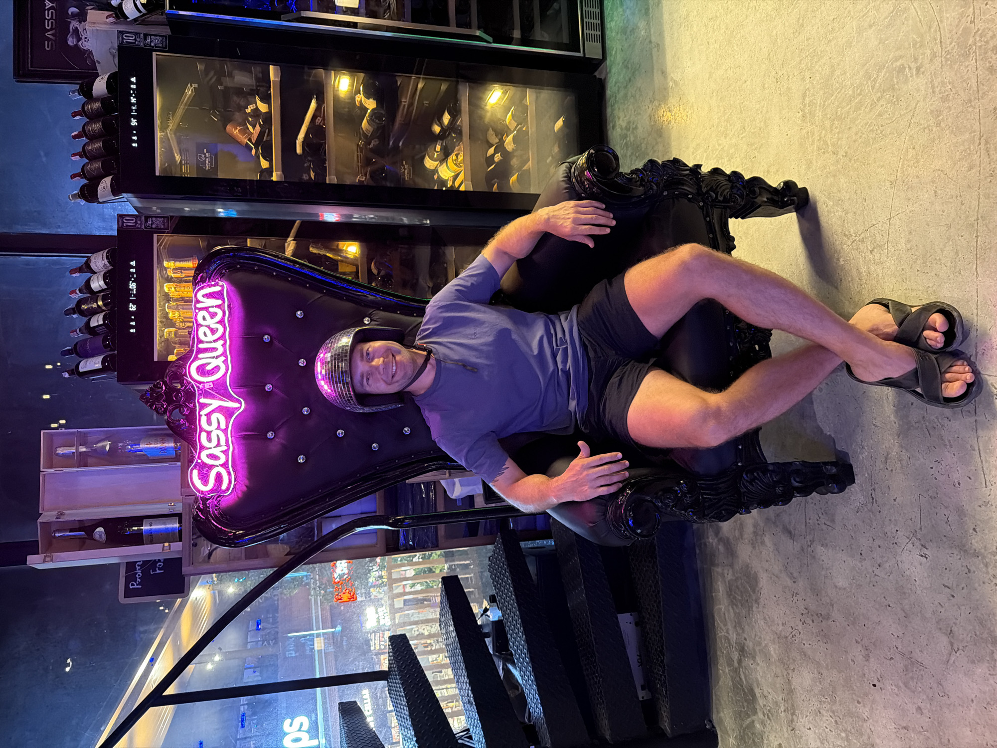 A man with short blond hair, wearing a silver disco ball helmet, a light blue t-shirt, and dark shorts, is sitting in a black throne-like chair. He is wearing dark gray sandals. The chair is adorned with a neon sign that reads "Sassy Queen." Behind the chair, a wine refrigerator is visible, stocked with bottles of wine. To the left of the chair, bottles of wine are displayed on shelves, along with a sign reading "Pro From Fa" (partially obscured). The scene appears to be inside a bar or lounge. The background shows a blurred view of the outside, seemingly a street with shops and lights, suggesting an urban environment. This photograph was taken in Baan Chaweng, Thailand.