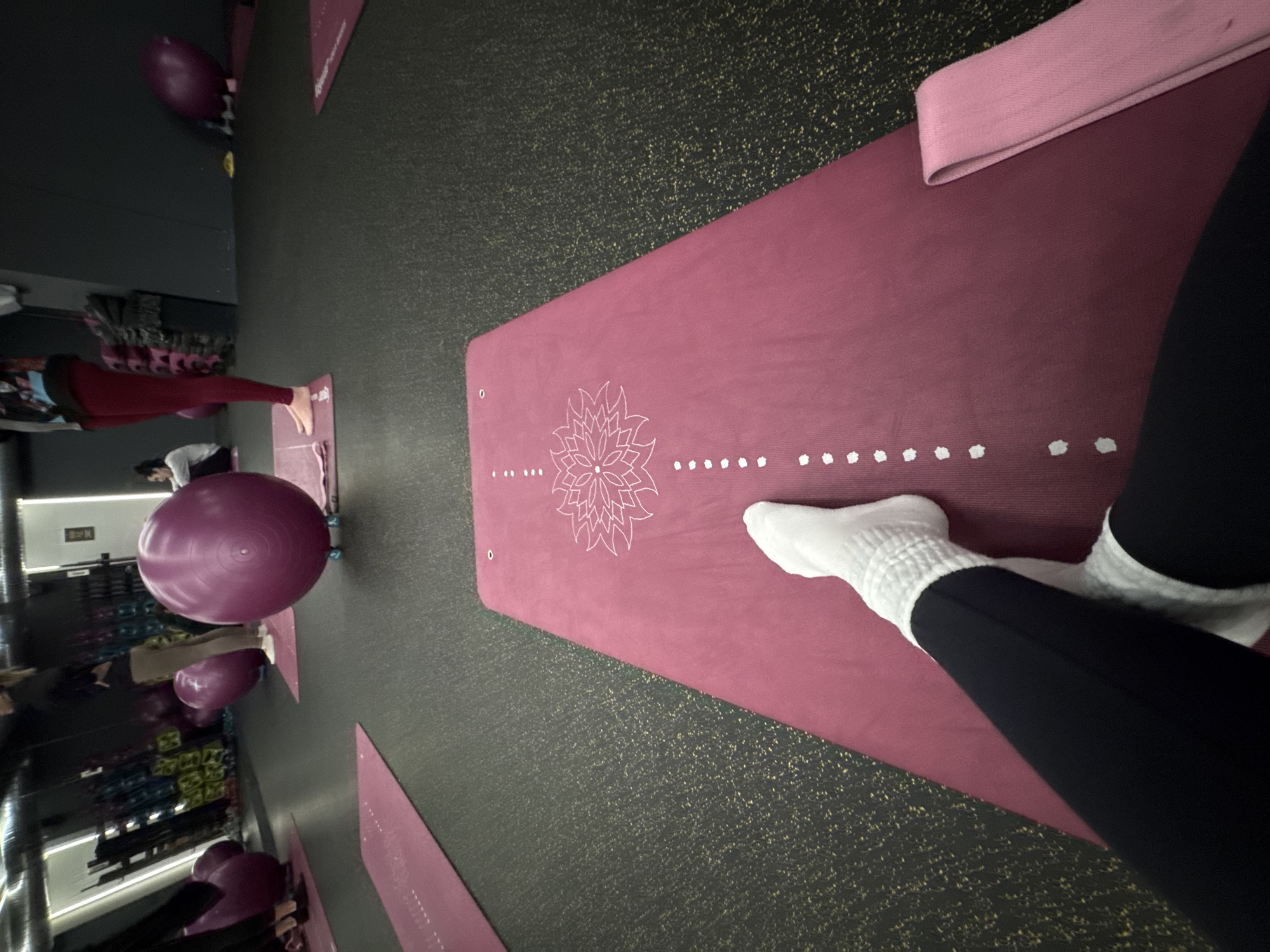 The image shows the interior of a fitness studio or gym, likely in Sofia, Bulgaria, with yoga mats and exercise balls visible. Several people are engaged in physical activities, possibly yoga or fitness classes. The foreground focuses on a pink yoga mat with a white lotus flower design and white dots along the edge. A person wearing black leggings and white socks is positioned on the mat. A pink resistance band is placed at the top of the mat. The studio has a dark floor and visible equipment and mirrors, indicative of a fitness or wellness center.