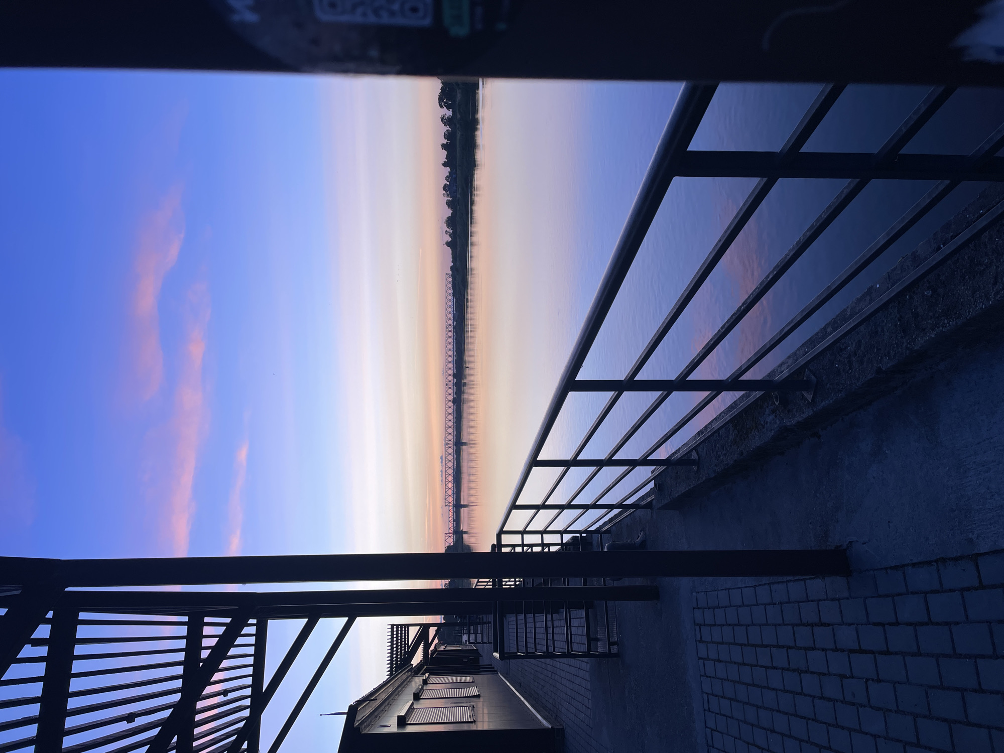 The image depicts a twilight scene by a river in Sovetsk, Russia, likely near the railway bridge that spans the Neman River. In the foreground, a dark metal railing runs along a walkway paved with dark bricks and concrete. To the left, a dark building with closed shutters and a slatted roof structure is visible. The river itself is calm and reflects the soft colors of the sky, which transitions from a deep blue at the top to hues of pink, orange, and light yellow near the horizon. A silhouette of a long railway bridge with intricate metalwork is visible across the river, and beyond it, a treeline marks the opposite bank. The overall atmosphere is peaceful and serene, characteristic of a sunset or sunrise.