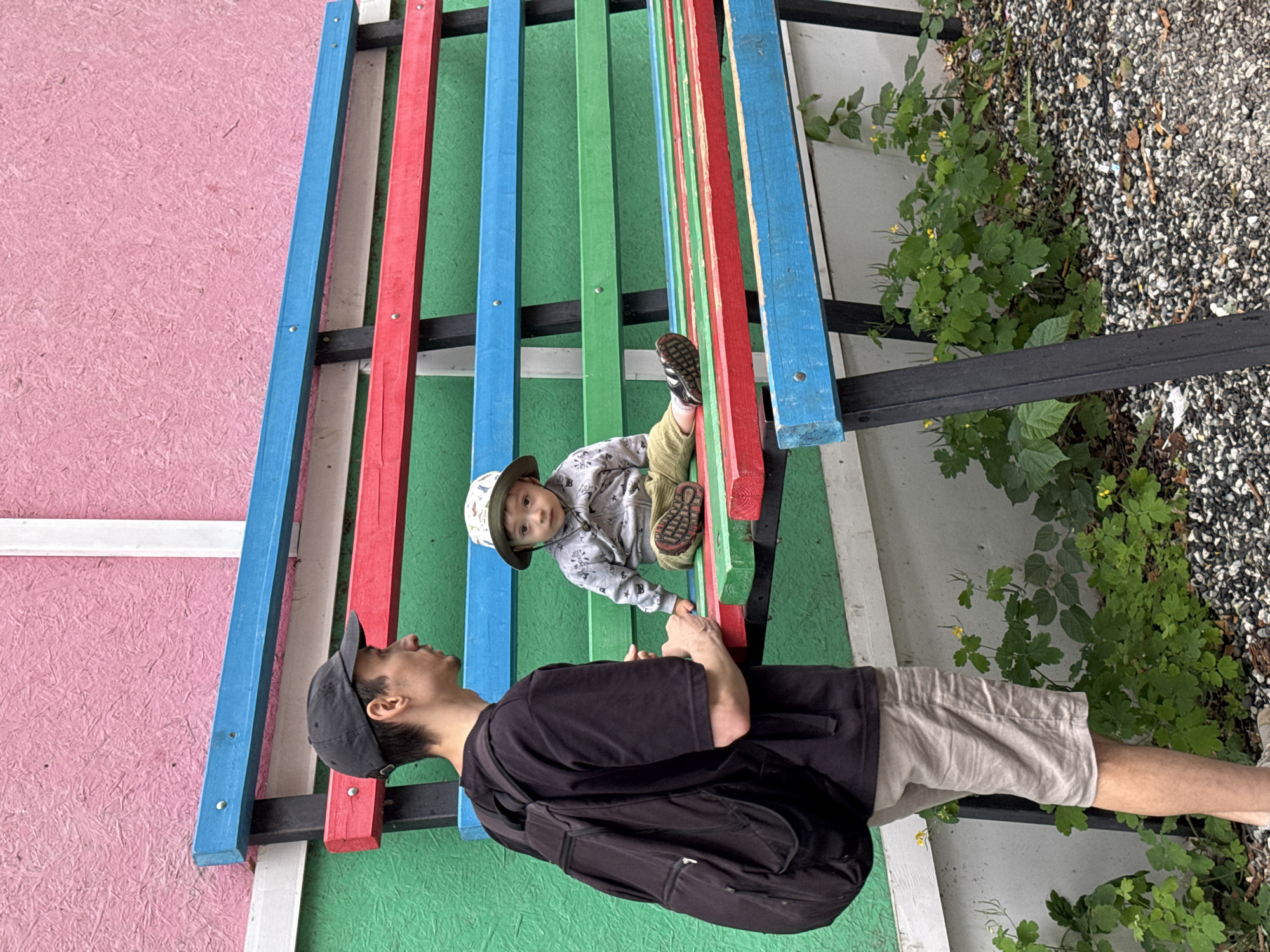 A man wearing a black t-shirt, a black baseball cap, and beige shorts, with a black backpack on his back, stands to the left of a colorful, multi-tiered bench. He is looking towards a child seated on the bench. The child, wearing a hat, a grey patterned hooded jacket, and light-colored pants, is looking towards the camera. The bench is painted with alternating red, blue, and green planks. Behind the bench, there is a wall painted pink on the upper portion and green on the lower portion. The ground in front of the bench is covered with small grey and white pebbles and some green vegetation with small yellow flowers. The overall setting appears to be an outdoor public space. There is no information in the image to suggest a specific location within the city of Izhevsk, Russia.