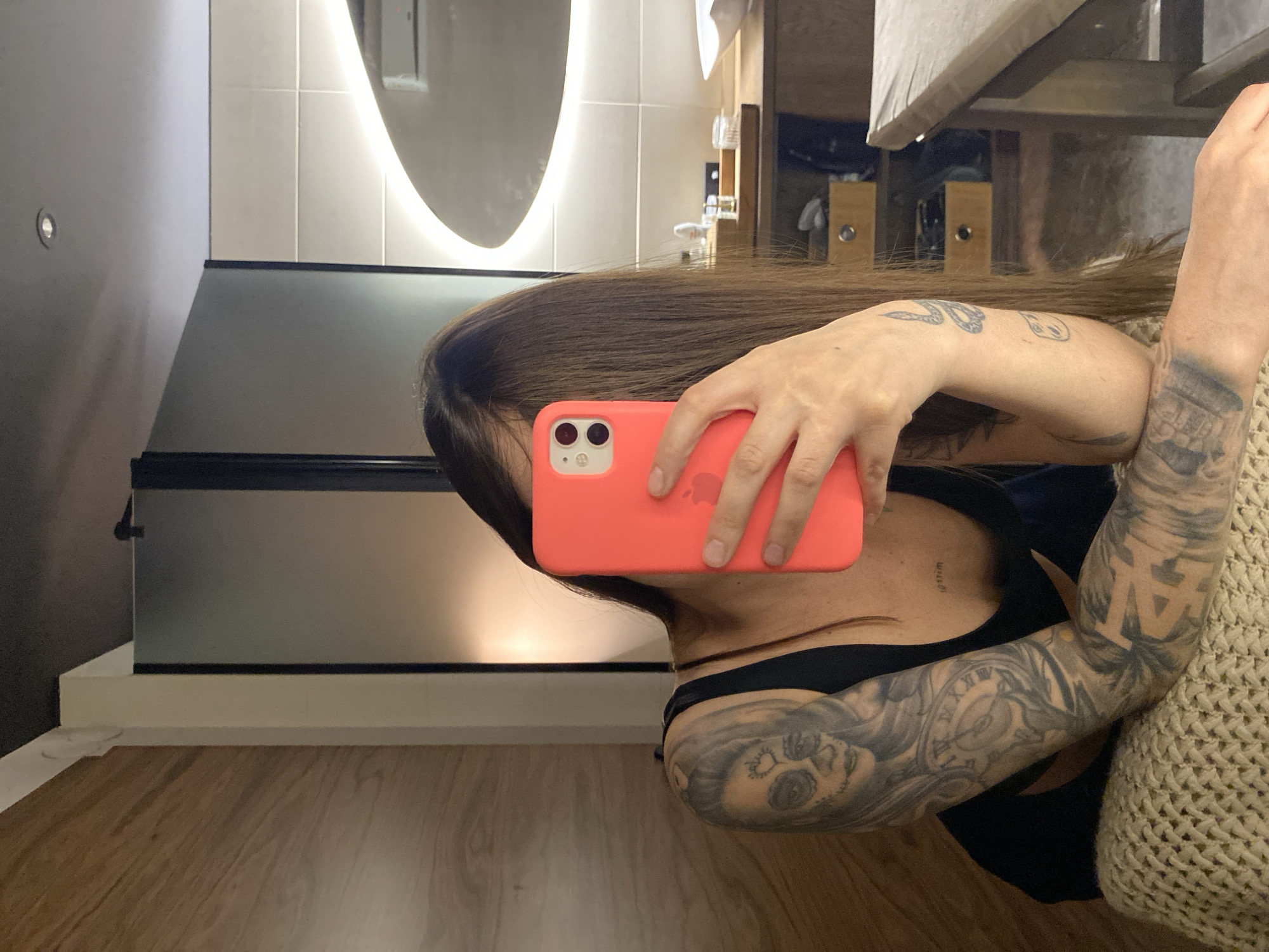 A person is captured taking a mirror selfie in what appears to be a modern bathroom, set in Votkinsk, Russia. The image is oriented vertically, showing the individual from the chest up, with their face obscured by long, dark hair and a coral-colored smartphone case. The person is holding an iPhone with their left hand, reflecting the back of the phone which displays a partially visible Apple logo. They are wearing a black top or bra, and a light-colored, possibly knitted, garment or blanket is draped over their right side.
Both arms are extensively covered in detailed tattoos, including a portrait-like figure near the left elbow, a snake design, and various abstract or geometric patterns on the right arm.
The bathroom features an oval or irregularly shaped mirror with a prominent LED backlight that casts a soft glow. Below the mirror is a sleek, dark-colored vanity or countertop. The walls consist of light-colored tiles directly behind the mirror and a contrasting darker, plain wall to the left, which has a small circular light fixture. The floor is covered with light brown wooden planks. The predominant lighting comes from the mirror's backlight and the small wall light, suggesting the photo was taken indoors, possibly in the evening or at night. Faint, indistinct text or numbers are barely visible on the person's chest/shoulder area, partially obscured by their hair.