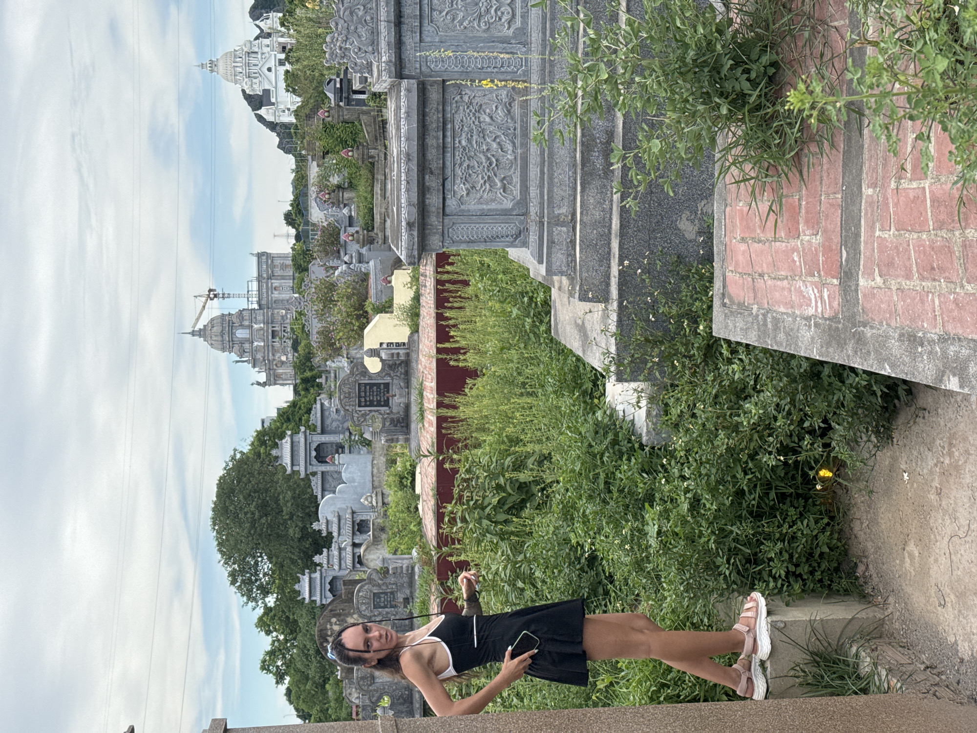 In Hoa Lư Ward, Vietnam, a young woman stands on a stone structure in a cemetery. She is wearing a black dress with white trim and pink sandals. She holds a black mobile phone in her right hand. In the background, several ornate tombs and mausoleums are visible, some adorned with intricate carvings. Beyond the cemetery, large, elaborate white buildings with domes, reminiscent of classical architecture, dominate the skyline, one of which appears to be under construction with a visible crane. The sky is overcast.