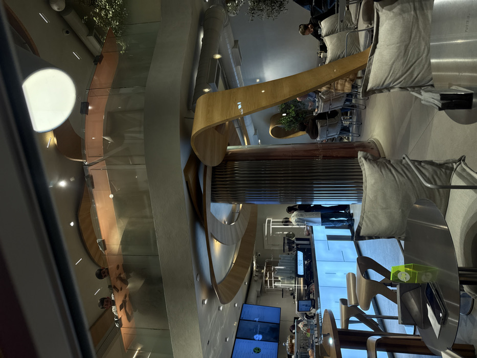 Upside-down shot of a modern restaurant. The space features curved architectural elements in wood and grey. Occupied chairs and tables are visible, along with customers and staff near a brightly lit counter area displaying menus. The decor is sleek, with metallic accents.