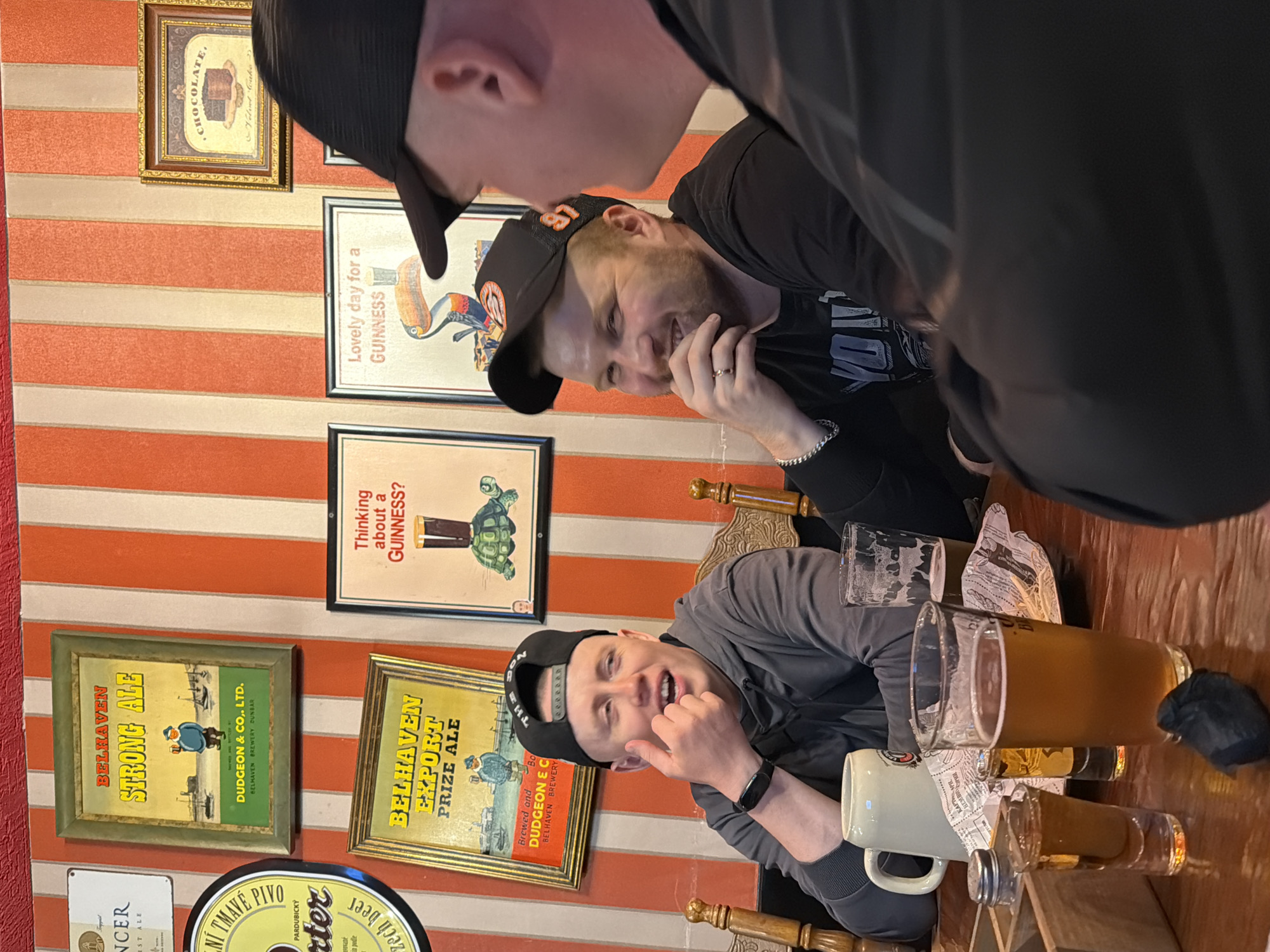 Three men are sitting at a wooden table with multiple beverages in what appears to be a pub or bar. The background has red and white striped wallpaper adorned with framed advertisements for alcoholic beverages, including Belhaven Strong Ale and Guinness. The men are interacting; one is primarily in the foreground, one is partially in the background wearing a baseball cap, and the third is mostly out of frame. There are glasses and a beer stein on the table. There is no information in the image to verify that the location is in the city of Chaikovsky, Russia.