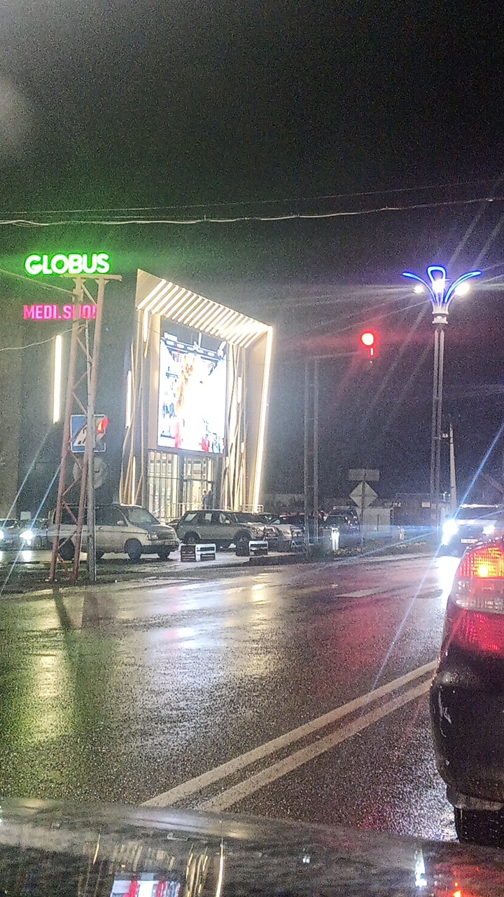 This media file captures a nighttime scene on a wet street, likely in Balykchy, Kyrgyzstan. The setting is an urban environment with a modern building facade illuminated by a large, bright LED screen. Above the entrance of this building, "GLOBUS" is prominently displayed in neon green, with "MEDI.SUO" visible below it in pink neon.
The street is wet and reflective, indicating it has recently rained or is currently raining. Cars are parked and driving on the street, their headlights and taillights creating streaks of light on the wet asphalt. One car in the foreground on the right side of the frame shows its red taillight.
A traffic light with a red signal is visible on a lamppost adorned with blue lights. The overall atmosphere suggests a busy commercial area at night, with the bright lights of the building and advertisements contrasting with the dark sky. The weather conditions and the wet road suggest a cool, damp evening. There are no distinct activities or events taking place, rather it appears to be a snapshot of everyday street life in a city at night during inclement weather. The mood is functional, highlighting the commercial and traffic elements of the scene.