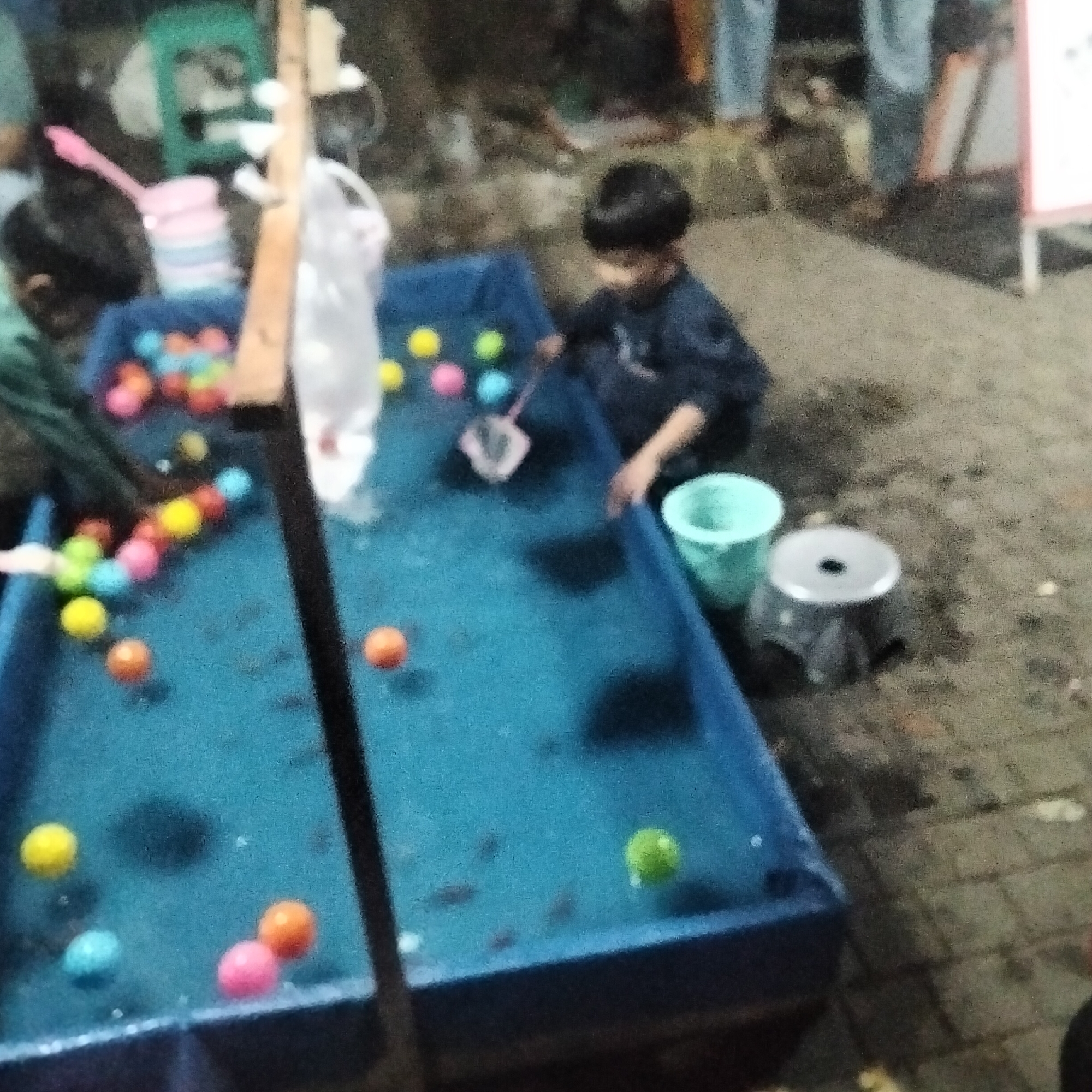 In Cisaat, Indonesia, a blurry, nighttime scene depicts a small fishing game. A young boy in a dark shirt is focused on the blue rectangular pool filled with water and small colorful balls. Next to him, a turquoise bucket and a silver stool are visible. In the background, other people are present but are out of focus. The overall impression is that of a lively outdoor gathering or market.