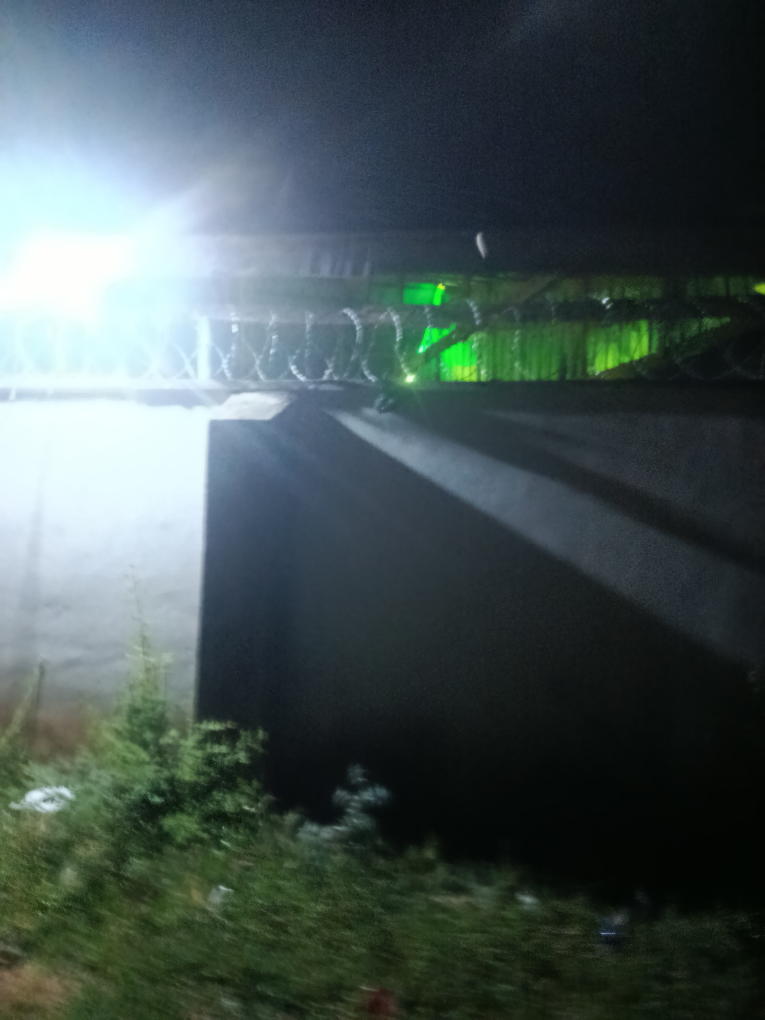 The image depicts a night scene with a bright light source illuminating a concrete wall and a barbed wire fence. The fence appears to be installed along the top of the wall. Behind the fence, there is a structure with green lighting, suggesting it might be a building or an area with illuminated signage. The ground in the foreground is covered with grass and some debris, indicating an outdoor environment. The overall impression is that of a secured perimeter at night. The location is described as being in Jalingo, Nigeria, which is a city located in the northeastern part of the country.