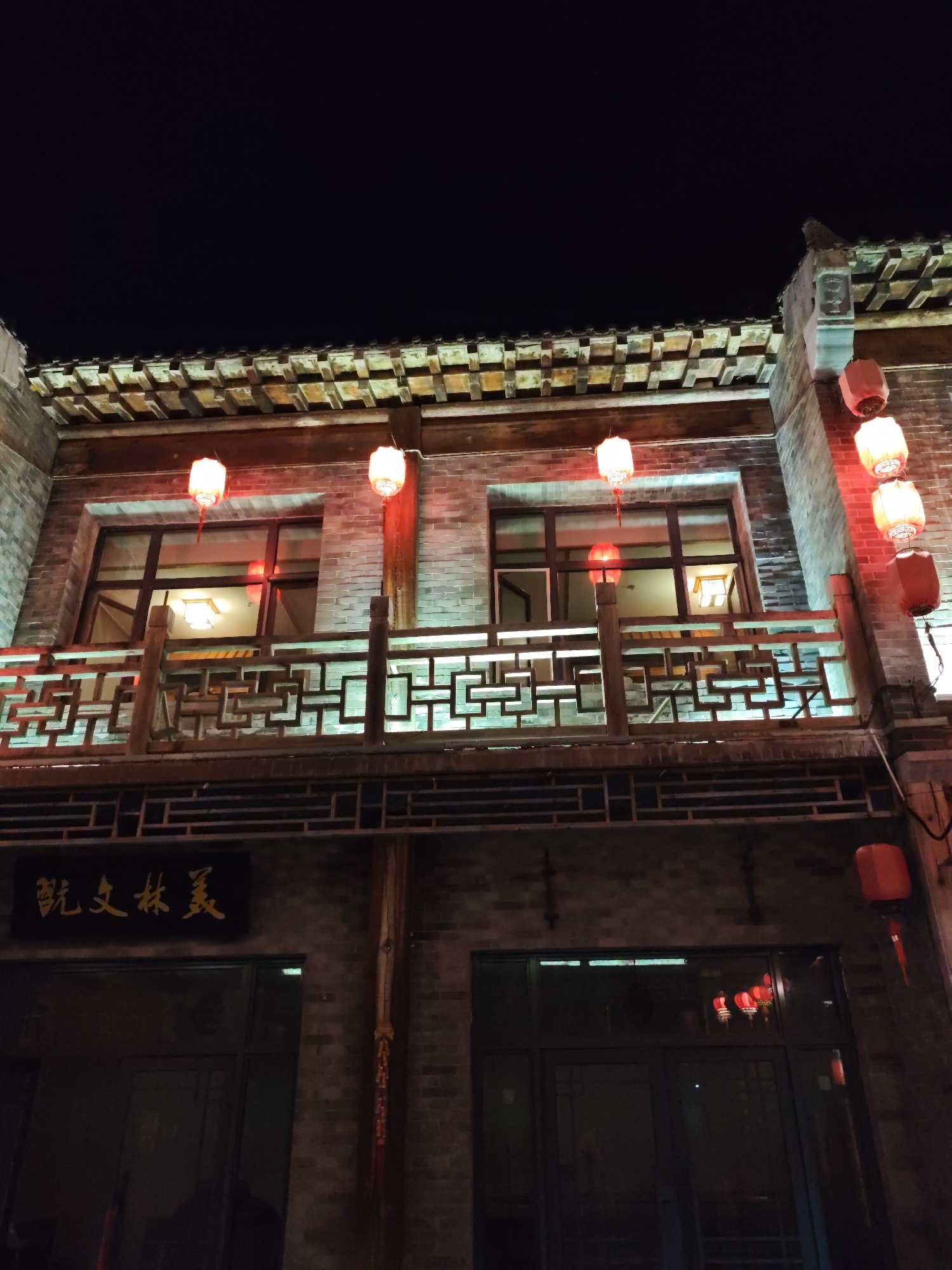 This is a night-time, low-angle photograph of traditional Chinese architecture in Lindian, China. The building features multiple levels with brick construction and a tiled roof. Red lanterns are prominently displayed, hanging from the eaves and along the facade, providing illumination. A wooden balcony with intricate lattice work spans across the upper level, with windows visible behind it. Below the balcony, a dark sign with gold Chinese characters is mounted on the brick wall. The ground level has large, dark-framed windows, reflecting some of the red lantern light. The overall impression is of an ancient building or a modern interpretation of traditional style, illuminated for nighttime ambiance.