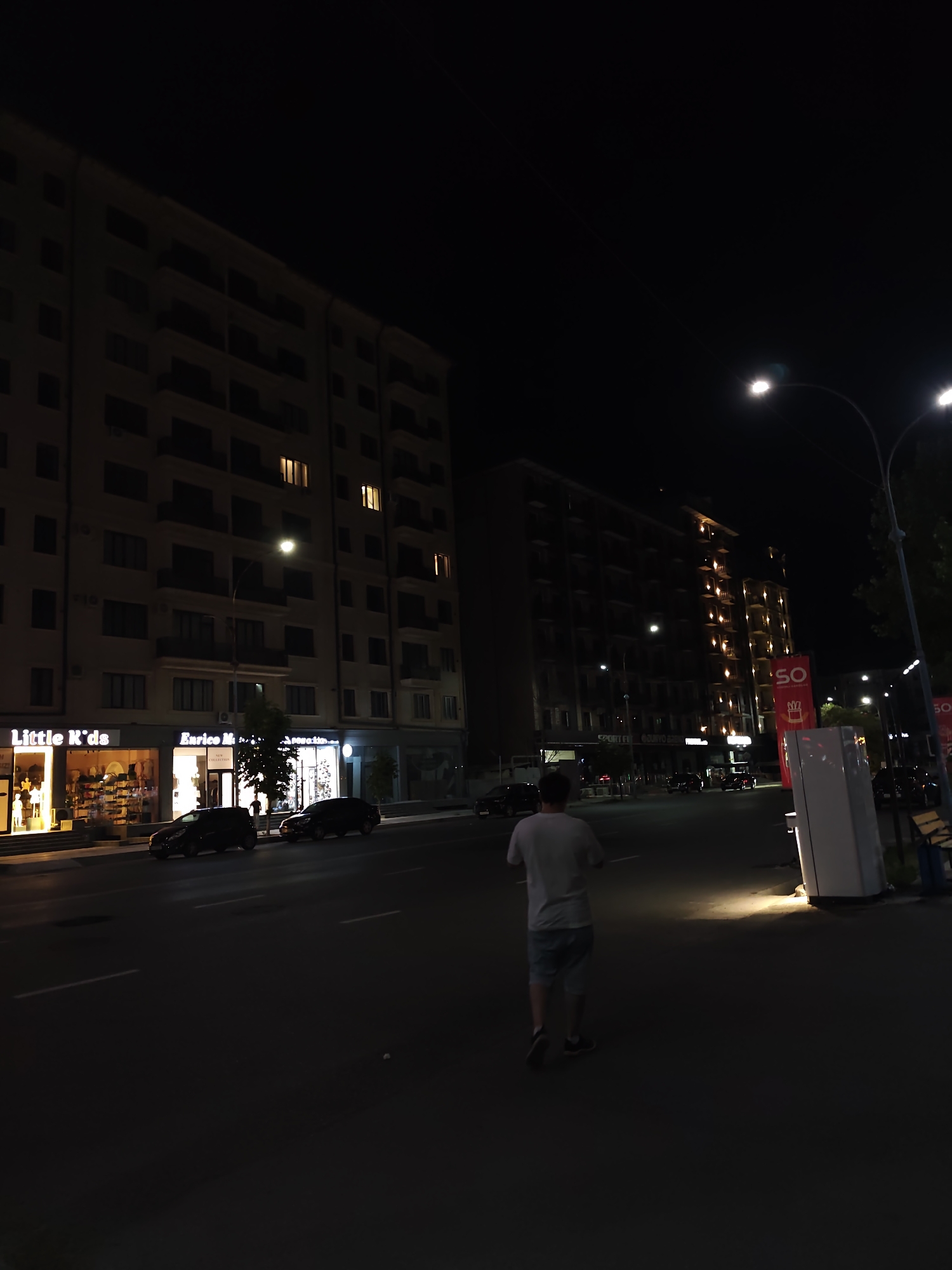 At night, a person walks on a sidewalk in front of buildings lining a street in Fergana City, Uzbekistan. The person, wearing a white t-shirt and light-colored shorts, walks away from the camera. On the left, storefronts are illuminated, with signs that read "Little K'ds" and "Enrico M." Dark-colored cars are parked on the side of the street. Streetlights illuminate the scene, casting light onto the pavement.