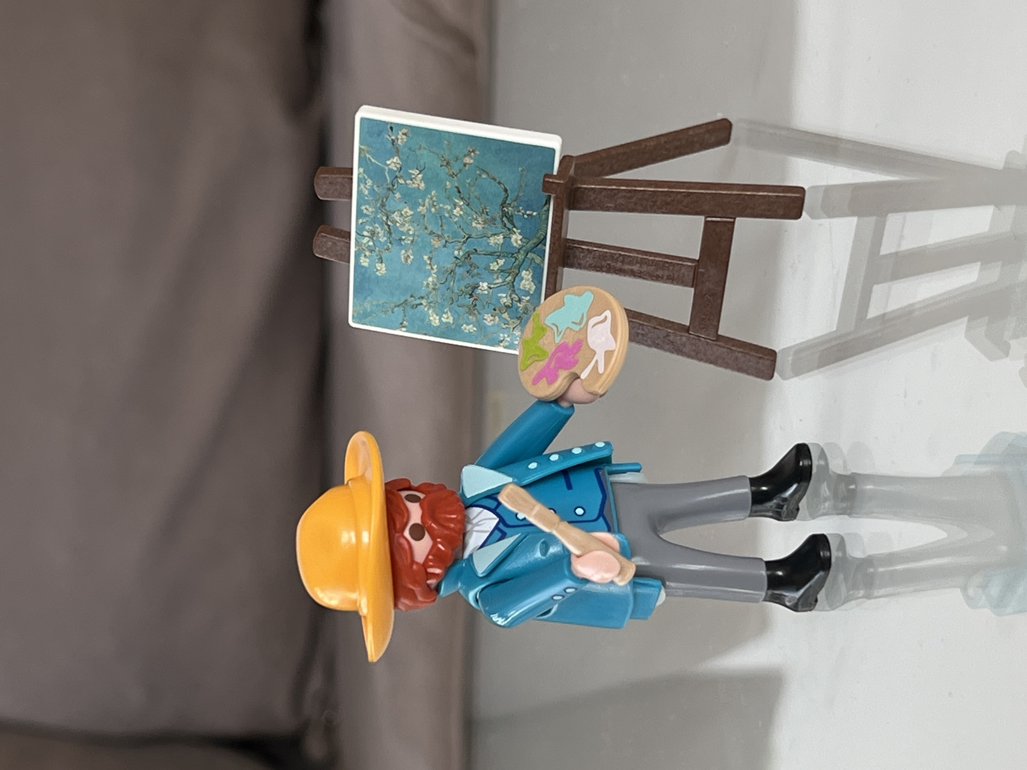 A Playmobil figure representing the artist Vincent van Gogh stands on a reflective surface, presumably a table. He holds a painter's palette in his right hand and a paintbrush in his left. The figure wears a yellow hat, a blue jacket, and grey pants with black boots. Behind him stands a brown easel displaying a miniature reproduction of Van Gogh's painting "Almond Blossoms." The background appears to be a blurred, neutral color. The location in the city of Bar, Montenegro, is not discernible from the image.