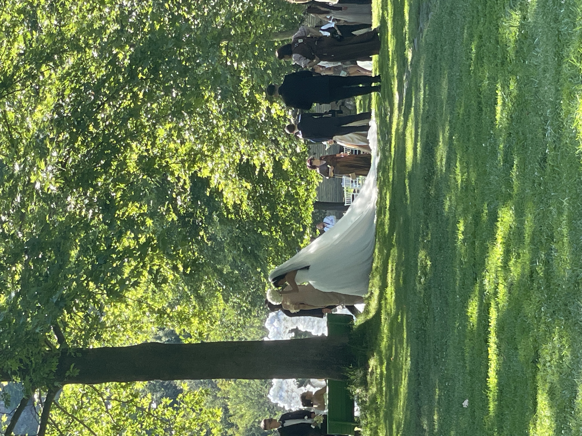 The image presents an outdoor event, consistent with a wedding, taking place in a verdant park setting in General Todorov, Bulgaria, under bright daylight. A dense canopy of green leaves from large trees covers the upper portion of the frame, casting dappled sunlight and shadow across the expansive grassy ground.
In the central mid-ground, a bride is visible in a white, strapless wedding gown with a long, sweeping train and a delicate veil. She is positioned with her back partially turned towards the viewer, her head slightly averted as if interacting with an individual standing closely beside her. This accompanying person, dressed in a light-colored garment, appears to be assisting the bride.
To the left, a man in a dark suit, white shirt, and bowtie stands near a rectangular green structure, possibly a bench or planter. Partially obscured behind this structure and the man, a woman is seated. Further left and behind, a large, abstract installation of voluminous white floral or fabric elements is prominent.
To the right of the central figures, a small group of attendees is present. Two men in dark suits are visible, one with his back to the camera. A woman in a dark, possibly brown, garment with an apron-style overlay also stands in this group, facing away from the viewer. Additional guests in various formal attire are visible further in the background, interspersed among the trees and natural foliage. The ground is consistently covered in green grass, interspersed with areas of bright sunlight and deep shadow.