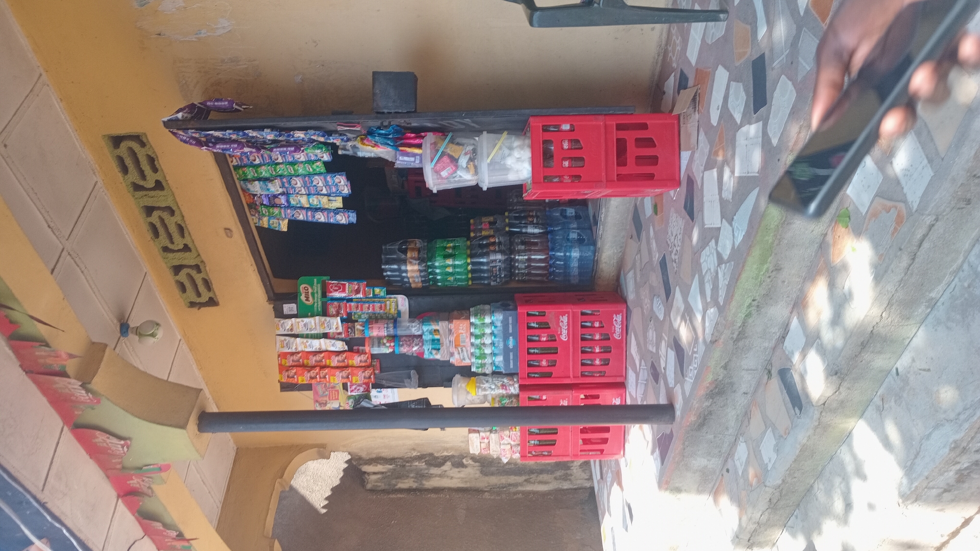 This image depicts a small roadside stall in Jalingo, Nigeria. The stall is filled with various goods, including packaged snacks, drinks in plastic bottles and crates, and what appear to be toiletries or personal care items. Prominently displayed are red Coca-Cola crates, some filled with bottles, and other crates holding water bottles. Shelves are stocked with colorful packets of what are likely snacks.
In the foreground, a person's hand is visible, holding a smartphone, suggesting a transaction or perhaps the person is taking a picture of the stall. The overall scene is of a typical commerce setting, likely in a busy or semi-busy area. The lighting suggests it is daytime, possibly late morning or afternoon, with shadows indicating sunlight. The ground is paved with mosaic-like tiles. The stall is constructed with a yellow painted facade, and the overall impression is one of a functional, local business. No specific emotions are discernible, but the scene conveys everyday activity. The visible text includes "Coca-Cola" on the red crates and "MILO" on a green package on one of the shelves.