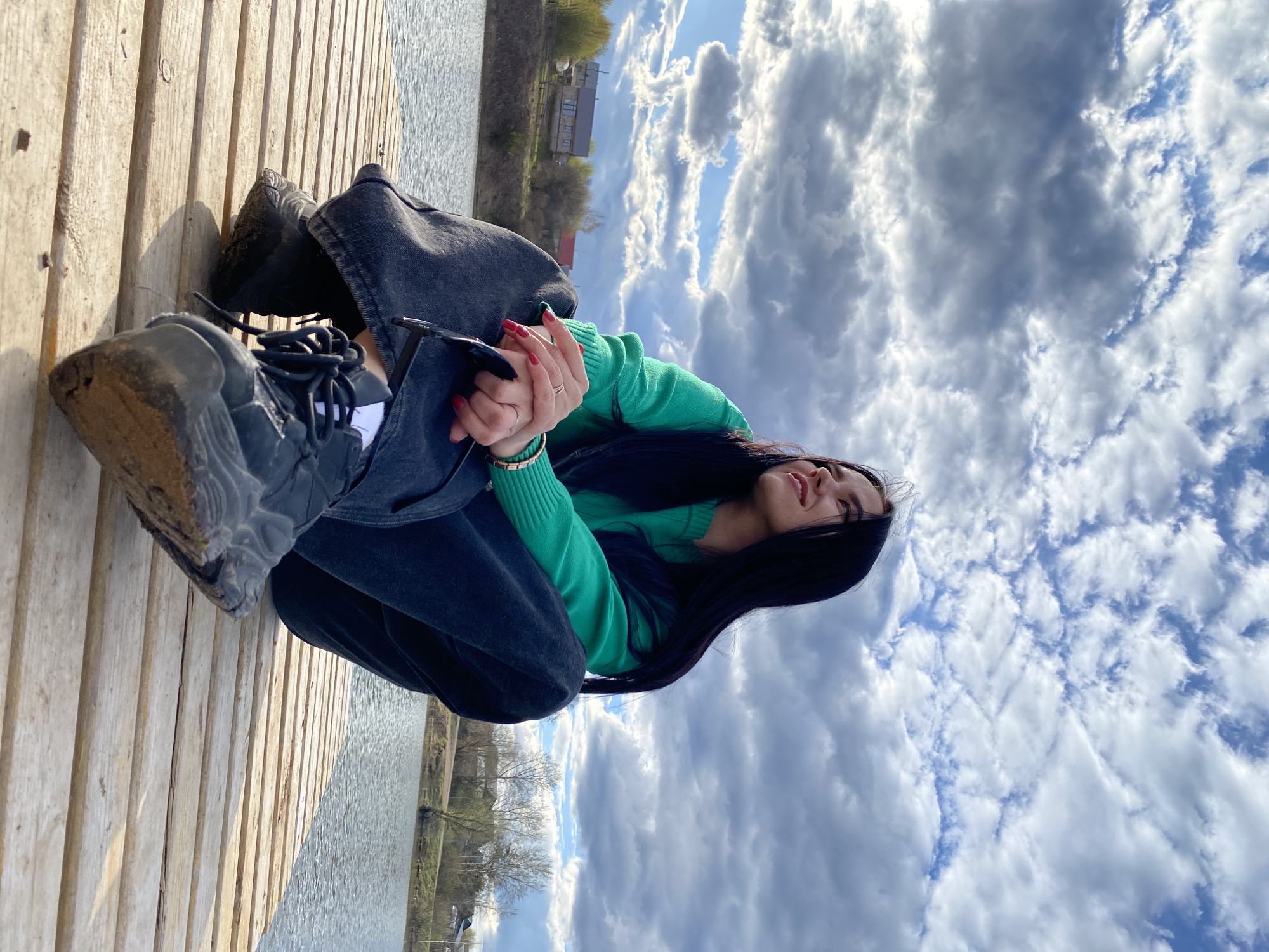 A young woman with long, dark hair is captured in a medium shot, sitting on a wooden dock next to a body of water. She is wearing a bright green long-sleeved sweater, dark jeans, and dark, chunky athletic shoes that appear well-worn. Her fingernails are painted red, and she wears a ring on her left hand, which is clasped lightly over her right. Sunglasses are tucked into her top near her shoulder.
The woman is looking upwards with a broad, joyful smile, conveying a sense of contentment and relaxation. The setting is outdoors, likely a river or lakefront in Фоки, Russia. The wooden planks of the dock are visible beneath her, leading towards the water. The water reflects the daylight, suggesting a bright day. In the background, across the water, there are trees (some still bare, others showing early green leaves) and distant structures. Above her, the sky is expansive and filled with a mixture of bright white and grey clouds, with patches of clear blue sky visible. The weather appears partly cloudy but bright, suggesting late morning or afternoon. There is no visible text in the image.