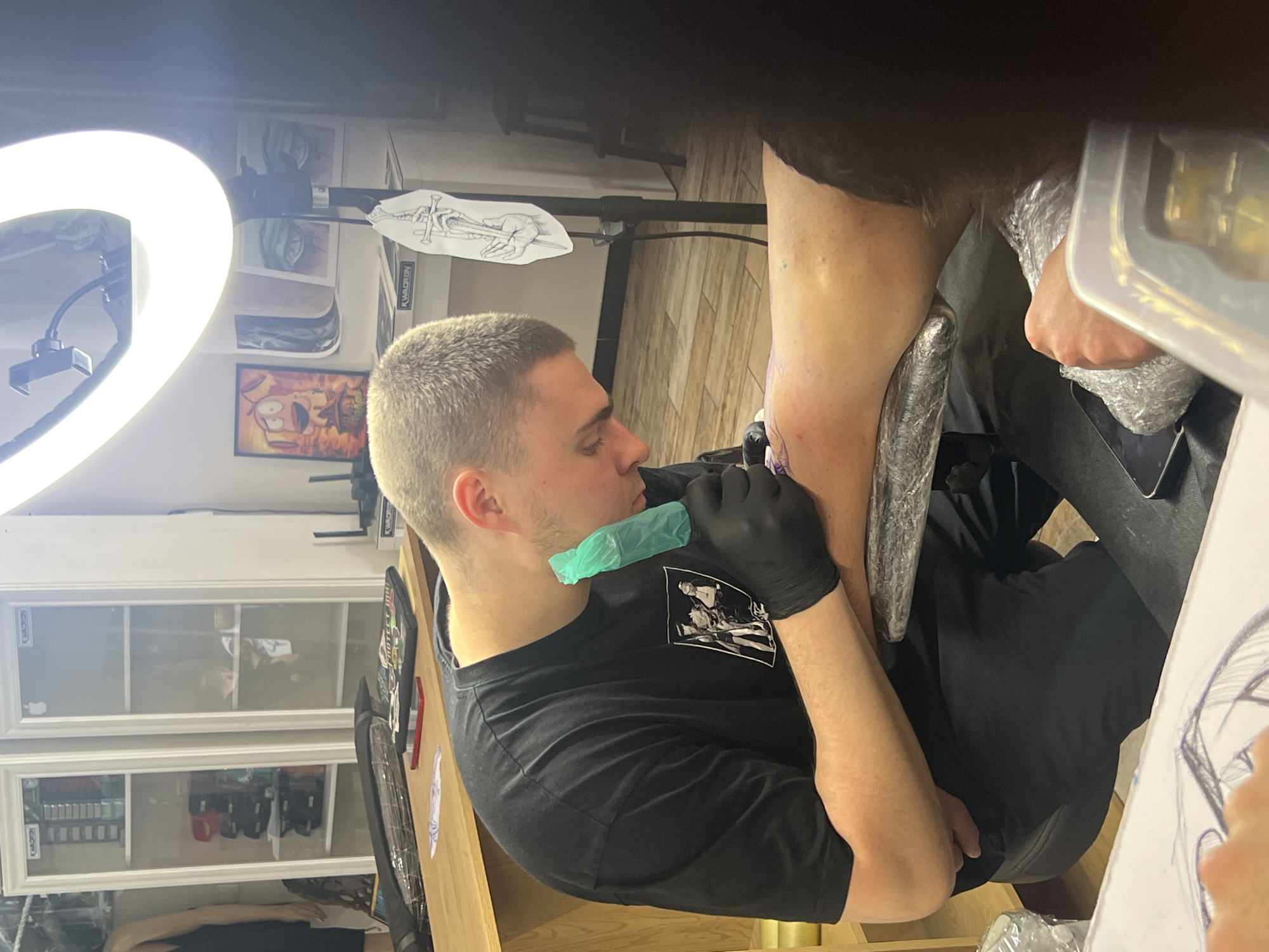 The image shows a tattoo artist in Sofia, Bulgaria, tattooing a client's arm. The artist, a fair-skinned man with short hair, wears a black t-shirt, black gloves, and a green mouth guard. He is focused on his work, holding a tattoo machine in his gloved hand. The client's arm is visible, wrapped in what appears to be a protective covering, with the beginnings of a tattoo visible.
The setting seems to be a tattoo studio. There is a ring light providing illumination. In the background, tattoo-related artwork is visible on the walls. Cabinets with tattoo equipment and supplies are also visible. A piece of paper with a drawing sits on a table.