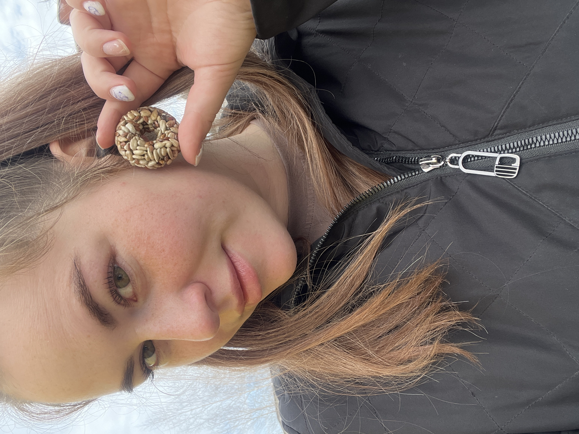 An outdoor close-up portrait in Chaikovsky, Russia, features an individual, likely female, with light brown hair, green or hazel eyes, and freckles on her face. She looks directly at the camera with a slight smile. Her right hand, featuring manicured nails with a light polish and faint floral designs, holds a ring-shaped object composed of various seeds, including sunflower seeds, near her right ear. She is wearing a dark, quilted jacket with a visible zipper that has a stylized metallic safety pin-shaped pull. A light brown garment is visible beneath the jacket at the neck. The background is out of focus, showing an overcast sky and the silhouettes of bare tree branches.