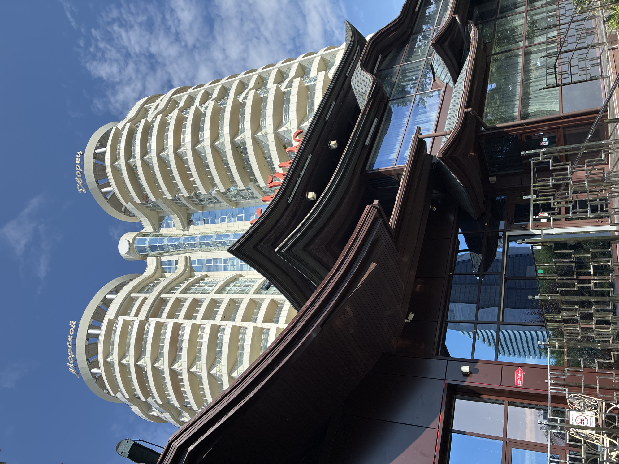 The image, taken from a low angle, captures a modern, multi-story building with a distinctive curved facade and numerous balconies. The building's top is adorned with text in Cyrillic, "Морской" on the left tower and "Дворец" on the right, indicating "Seaside" and "Palace" respectively. In the foreground, a building with dark brown, tiered roofing and large glass windows obscures part of the larger structure. Reflections of the curved building are visible in the windows of the foreground structure. The sky above is a clear blue with scattered white clouds. A camera is mounted on the exterior of the foreground building, and signage, including a red arrow and text that reads "Кофейня" (Cafe), is visible near an entrance. The overall scene appears to be a commercial area or resort in Sochi, Russia, given the context of the Cyrillic text and architectural style.