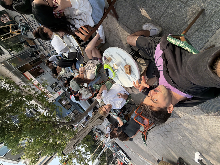 The scene captures an outdoor cafe on a paved sidewalk alongside a commercial street in Tel-Aviv, Israel. Daylight illuminates the area, casting distinct shadows. In the immediate foreground, a male with dark hair, wearing a black zipper jacket over a purple shirt and black shorts, sits on a green-patterned chair. He faces the camera directly, displaying an expressive facial gesture with widened eyes and slightly open mouth, while holding both hands up with splayed fingers. His right leg is extended, revealing a white sneaker.
Adjacent to him, a male in a white t-shirt and light-colored pants is seated at a round white marble-patterned table, holding a white cup and a smartphone, oriented towards the foreground subject. His table holds a sandwich, two coffee cups, and a bright green object.
Further into the mid-ground and background, approximately ten additional individuals are seated at outdoor tables. These include a female in a beige knit sweater drinking from a glass, a male in a black t-shirt utilizing a laptop, and a female in a white top and blue shorts engaging with a phone. The tables are various small, round cafe models, accompanied by chairs of different styles (green patterned, orange, wooden).
An open beige outdoor umbrella provides shade over a section of the seating. Behind the cafe, multi-story buildings line the street, featuring ground-level shopfronts. Visible signage includes Hebrew text, with identifiable English text for "BURGER SALOON" and "TLV." A tree with green leaves is situated near the cafe. The adjacent street shows parked vehicles, including a white car, and a scooter is partially visible on the sidewalk.