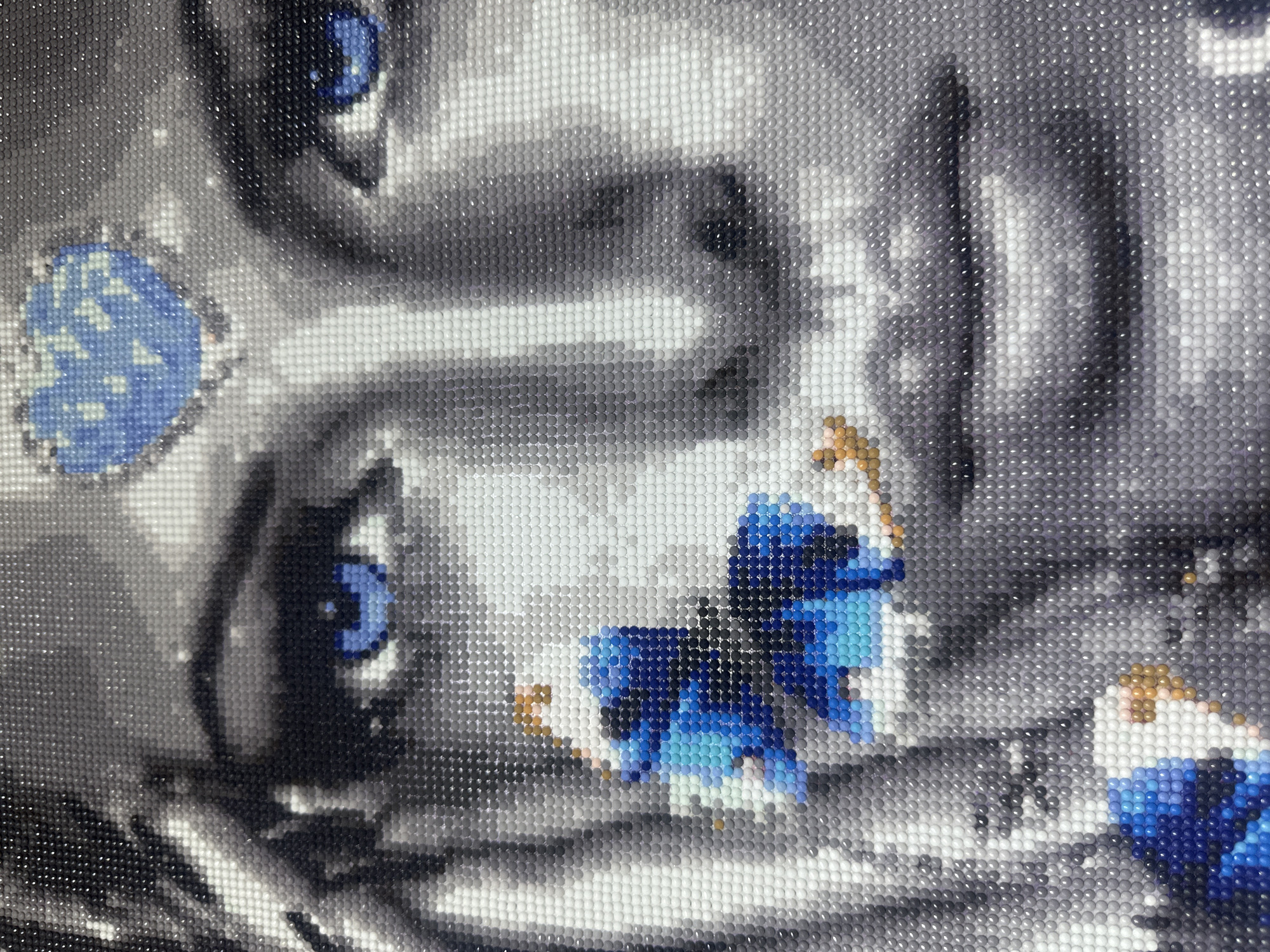 The image is a close-up of a diamond painting depicting the face of a woman. Her eyes are a striking blue, and there are blue and light blue diamond accents on her cheek, resembling a butterfly or floral design. A blue, heart-shaped diamond adorns her forehead. The rest of the painting is rendered in shades of grey and white, creating a monochromatic portrait. The diamond painting is created by adhering small, colored gems to a pre-printed canvas, forming a mosaic-like image. The location in Rudny, Kazakhstan is not discernible from the image itself.