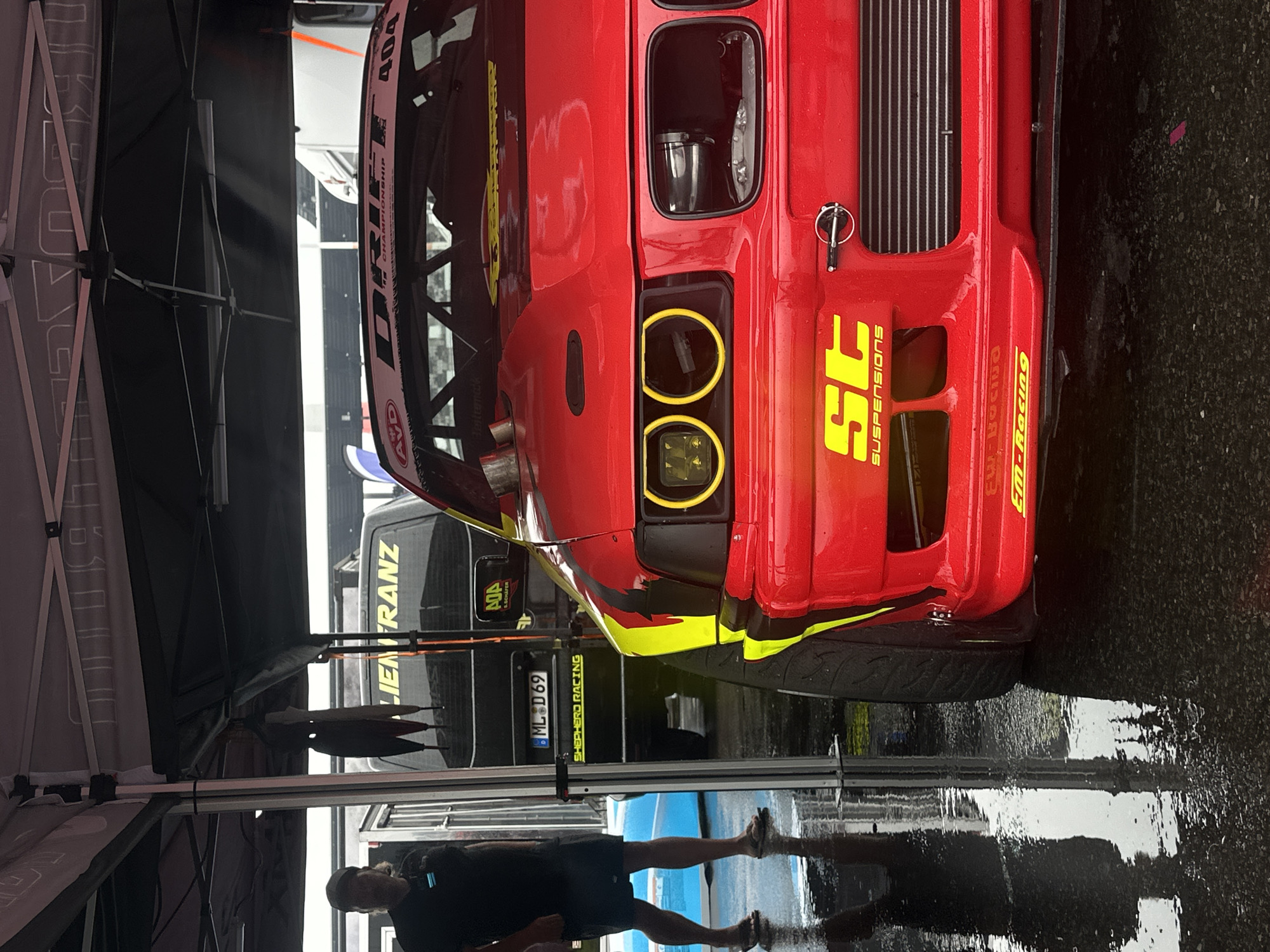 The image depicts a red drift car with yellow accents parked under a tent at the Lausitzring race track in Schipkau, Germany. The car's front end, including its distinctive yellow-ringed headlights, is prominently featured. In the background, a black van with "LIENFRANZ" and "SCHEFER RACING" decals is visible. A person wearing a black t-shirt and shorts stands to the left of the van, facing away from the camera. The ground is wet, suggesting recent rain, and reflections of the tent and car are visible on the surface. The overall scene appears to be part of a motorsports event.