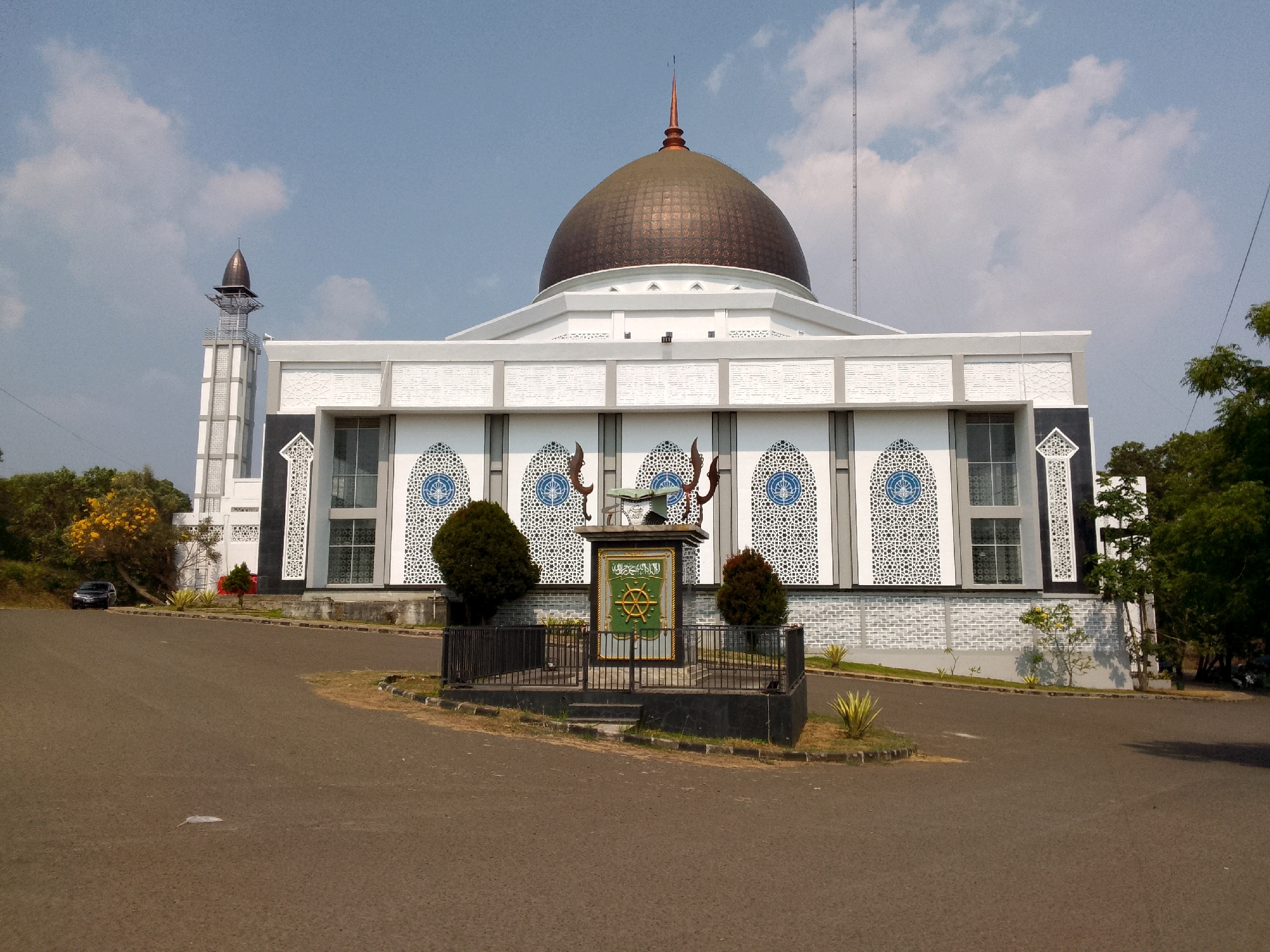 A wide, eye-level, exterior shot captures the front of a modern mosque, situated in a landscaped area with a paved road in the foreground. The mosque's architecture features a large, copper-colored dome crowning the main building, which is predominantly white with decorative gray accents and intricate white latticework above the windows. To the left, a tall, slender minaret with a pointed top rises, also featuring a similar copper-colored finial. In front of the mosque, a central monument or marker is visible, enclosed by a black metal fence. This marker displays a green emblem with golden Arabic calligraphy and a circular design at the top. The surrounding environment includes several green trees and bushes, with a small patch of yellow flowers on a tree to the far left. A dark-colored car is parked on the left side of the road. The sky above is clear blue with scattered white clouds, suggesting daylight conditions. The overall scene depicts a well-maintained religious building and its grounds under a bright sky.