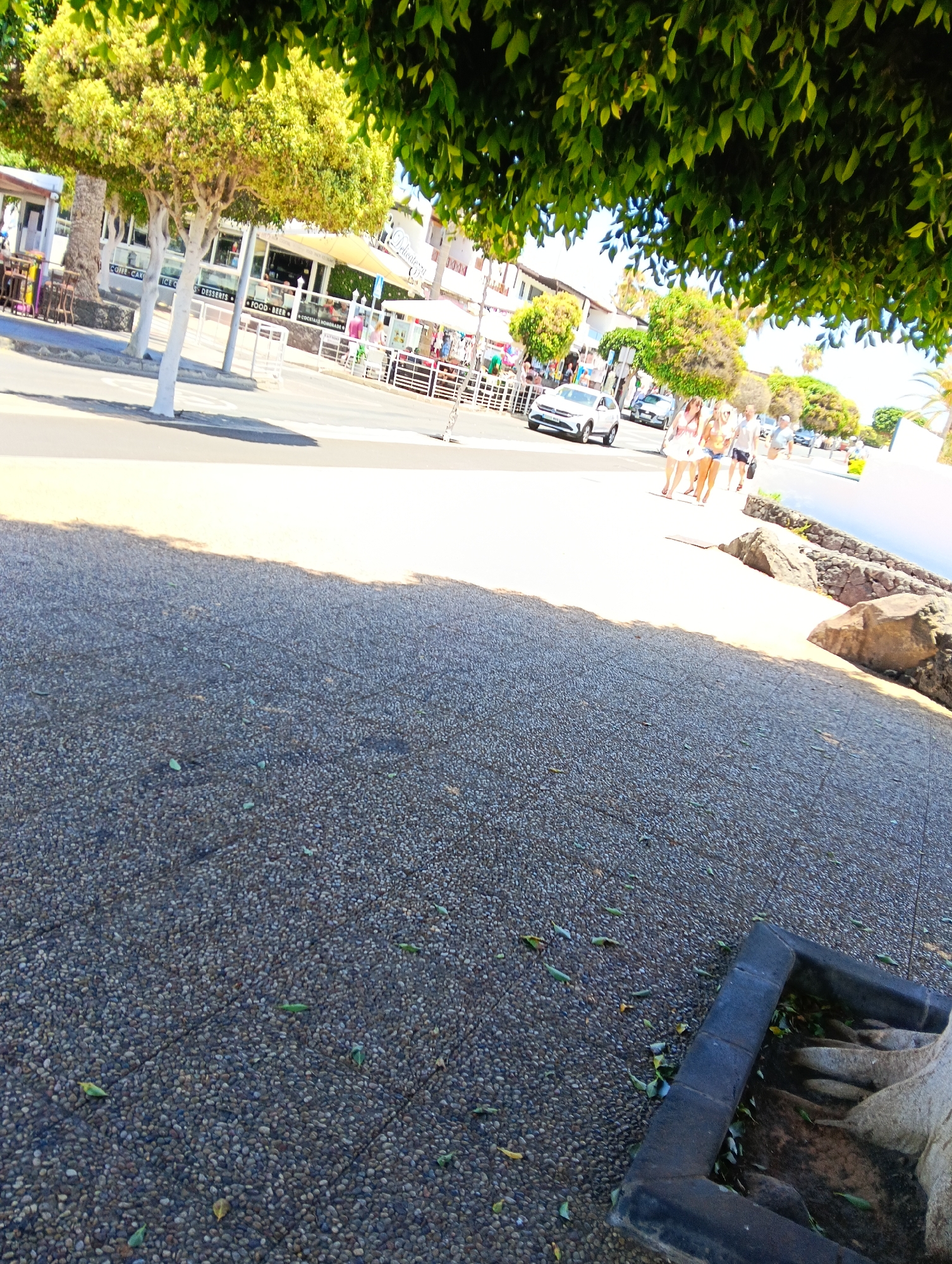 This vibrant image depicts a sunny daytime street scene in Puerto Del Carmen, Spain, showing a bustling commercial area. The foreground features a textured, pebbled sidewalk, partially shaded by the dense green foliage of overhead trees, casting distinct shadows. Along the right side of the path, large, rugged rocks are visible, suggesting proximity to a natural coastline or water body.
In the mid-ground, several people are walking casually along the sidewalk, dressed in light summer attire, indicating warm weather. A white SUV and another vehicle are seen driving on the road. Across the street, a line of shops and restaurants with outdoor seating, umbrellas, and awnings can be observed. Notably, some tree trunks in front of these businesses are painted white.
Visible text on the storefronts provides clues about the establishments, including "COFFEE CAKE," "ICE CREAM," "DESSERTS," "FOOD BEER," and "COCKTAILS HOMEMADE," with another sign reading "Delicacies." The strong sunlight and long shadows suggest it is late morning or early afternoon on a clear, bright day. The scene conveys a relaxed, lively atmosphere, typical of a popular resort town.