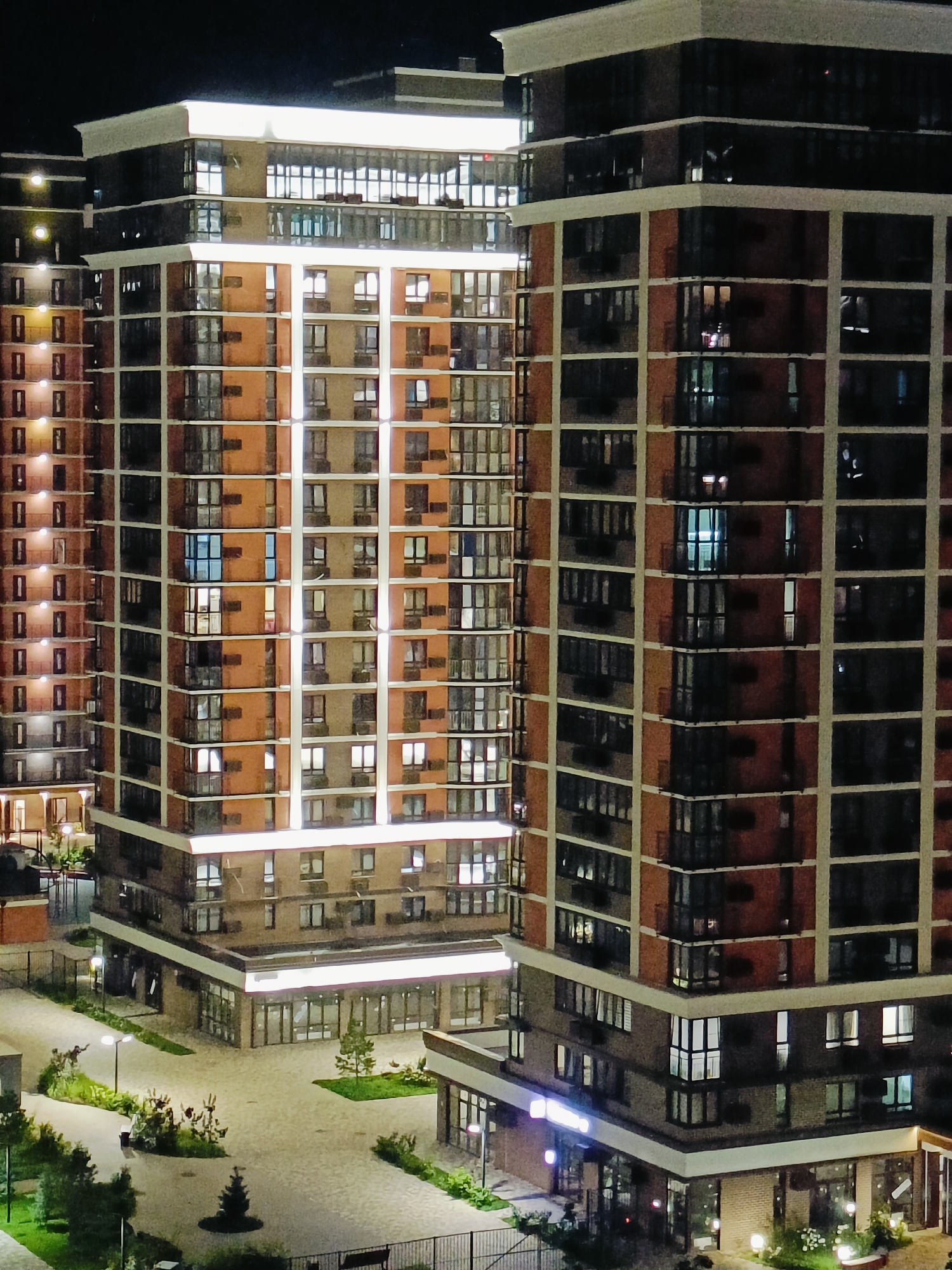 The image shows a night view of modern high-rise residential buildings in the city of Krasnodar, Russia. The buildings are characterized by their modular design, featuring a mix of brick and glass facades, with prominent vertical and horizontal white bands that are illuminated. Many windows are lit, suggesting occupancy. The ground level of the buildings appears to house commercial spaces with large glass windows. Surrounding the buildings are paved courtyards with streetlights, landscaping including trees and bushes, and some outdoor furniture like benches. The overall impression is one of a contemporary urban residential complex at night.