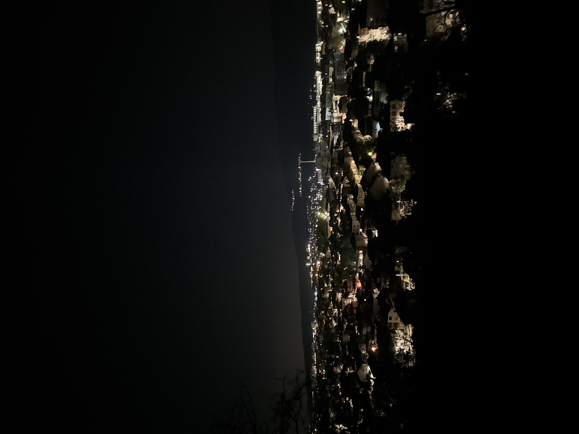 A nighttime view from Sveti Konstantin, Bulgaria shows an area of illuminated buildings and streetlights along the coastline. To the left of the image is a dark body of water, possibly the Black Sea. The lit-up area suggests a residential or commercial district with various buildings and structures, some with visible architectural details. The dark areas surrounding the lit-up zones likely represent undeveloped land, natural terrain, or shadows cast by buildings. The overall scene captures the urbanized and naturally dark environment of Sveti Konstantin at night.