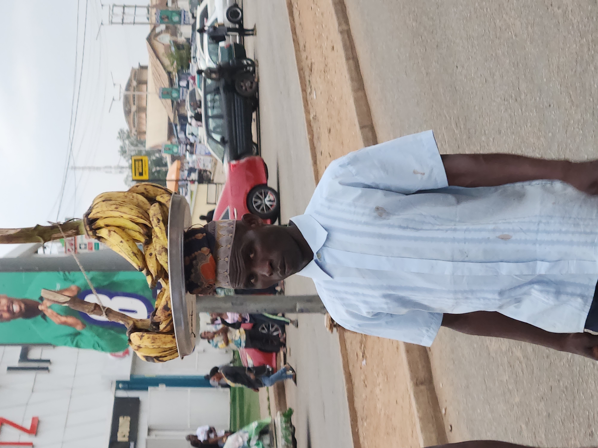 An eye-level outdoor medium shot shows a man carrying a tray of bananas on his head. He is located on a street, presumably in Jos, Nigeria. The man, who is dark-skinned, wears a light blue striped shirt and a head covering. The bananas are mostly ripe and arranged on a metal tray. The man is facing towards the camera with a neutral expression. The background shows a busy street scene with cars, buildings, and pedestrians. A large billboard is visible behind the man and bananas. The general environment appears to be a commercial area with a mix of vehicular and pedestrian traffic.