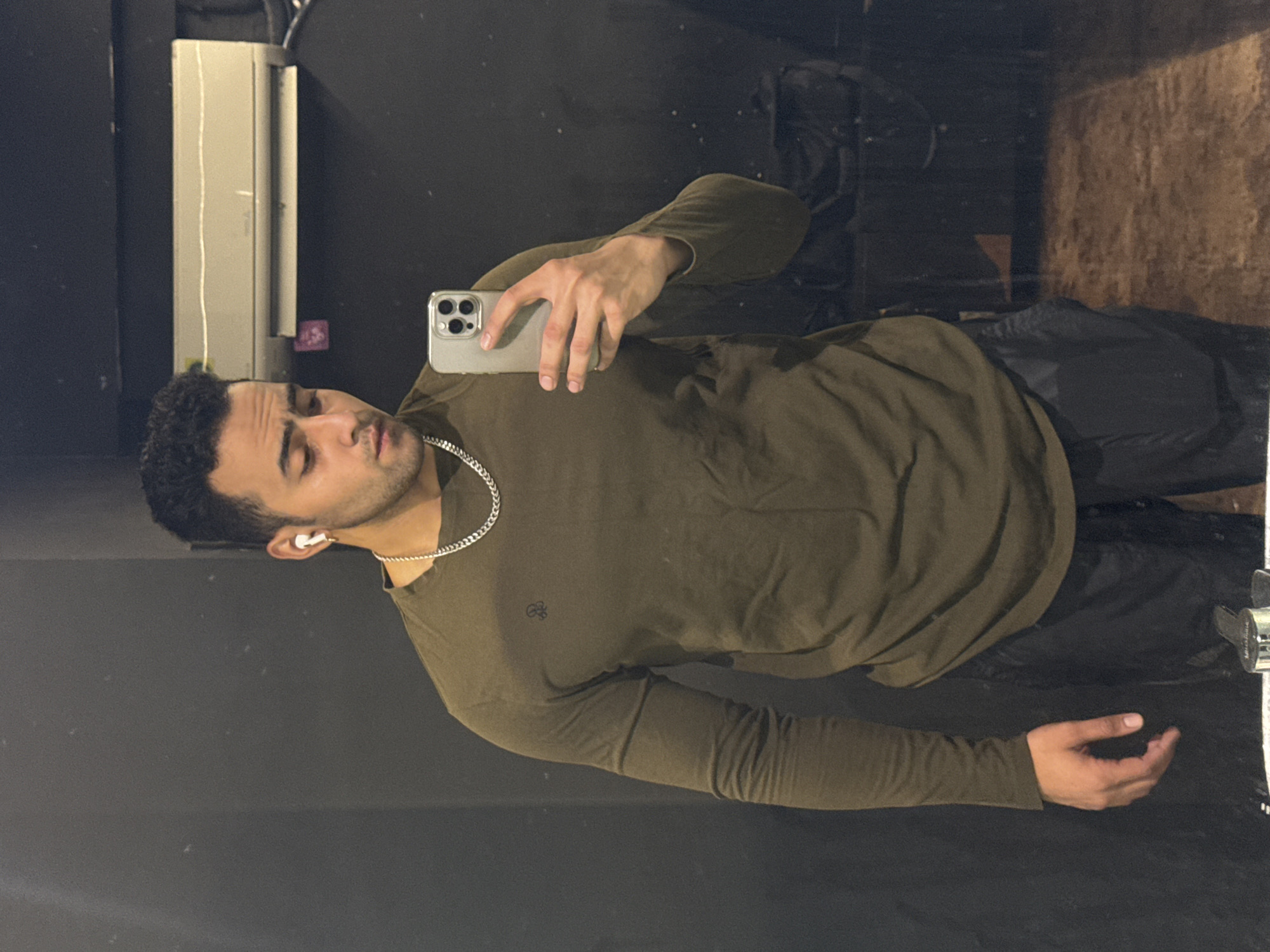 A male individual is captured in a mirror selfie within an indoor facility, likely a gym changing room or similar private space in Kolkata, India. The subject, a man with dark, short, curly hair and light brown skin, is positioned centrally, gazing downwards towards the smartphone he holds in his right hand. He wears a dark olive green long-sleeve t-shirt with a small dark emblem on the left chest and black athletic pants or shorts. A silver-colored chain necklace is visible around his neck, and white wireless earbuds are present in both ears. The smartphone, an iPhone with a light-colored case featuring a triple-lens camera array, is held in a selfie orientation.
The background consists primarily of a dark, reflective surface, suggesting a mirror or polished wall. In the upper right quadrant, a white wall-mounted split air conditioning unit is visible. Its main body is horizontal, with a white, ribbed top surface and a slightly darker intake vent below. Black electrical wiring extends from the unit's right side. Reflections on the dark surface indicate a dark duffel bag or backpack in the lower right background. At the bottom edge of the frame, a metallic fixture, possibly a faucet spout, is partially visible above what appears to be a countertop or sink basin. The overall lighting is subdued.