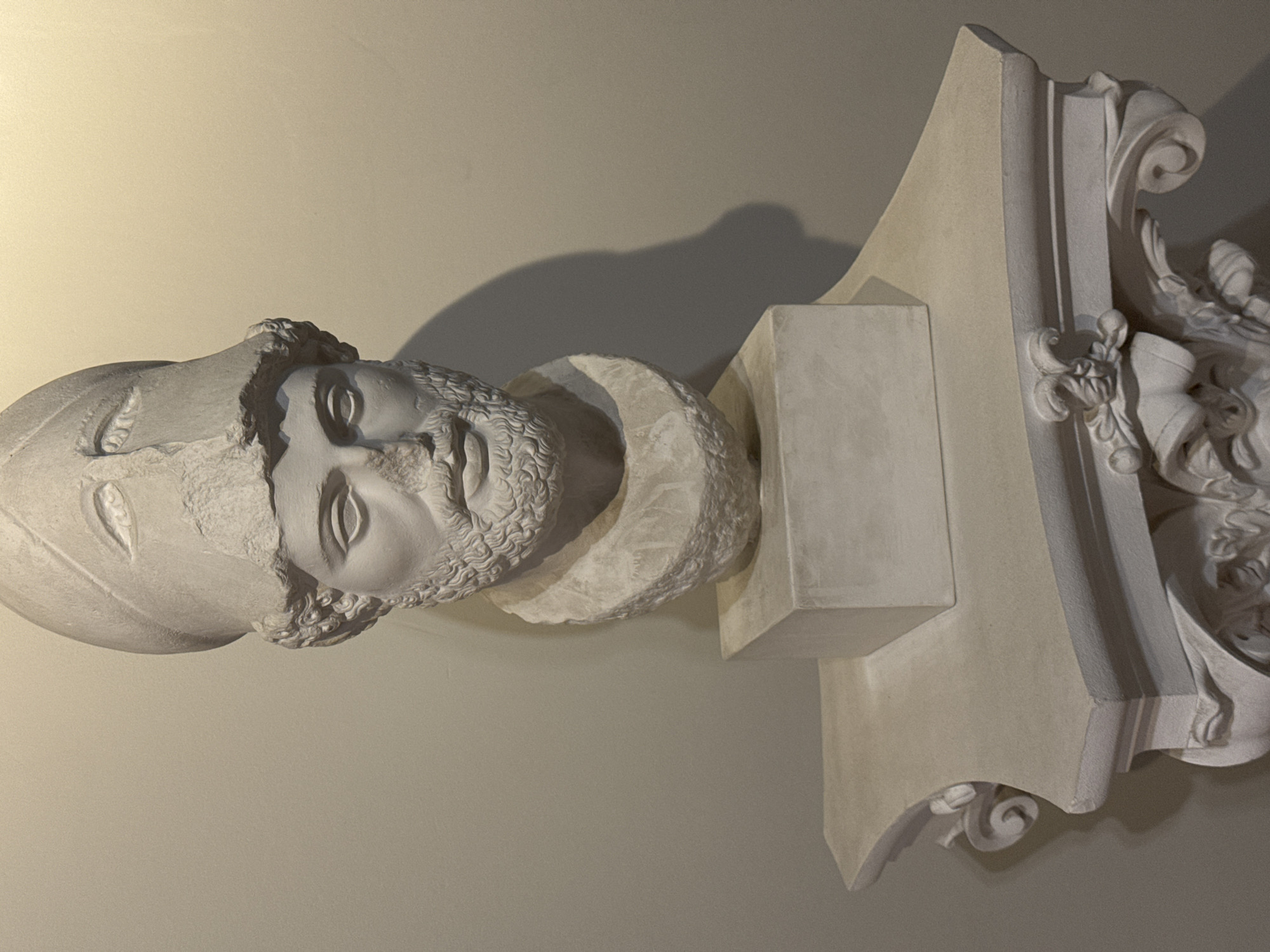 A bust of a man wearing a helmet and a beard rests on a square pedestal, which is placed on an ornate, white stone pedestal. The bust is made of white stone and appears to be a classical sculpture. The man has a stern expression and his eyes are looking forward. The helmet is adorned with what appear to be stylized eyebrows. The background is a plain, light-colored wall, with a shadow cast by the sculpture. The ornate pedestal has intricate carvings, including volutes and floral motifs, suggesting it is part of a larger architectural element or decorative display. The lighting is soft and directional, highlighting the textures and forms of the sculpture. The scene is static, with no discernible action taking place. The overall impression is one of historical or artistic significance. This object is located in the Hermitage Museum in Saint Petersburg, Russia.