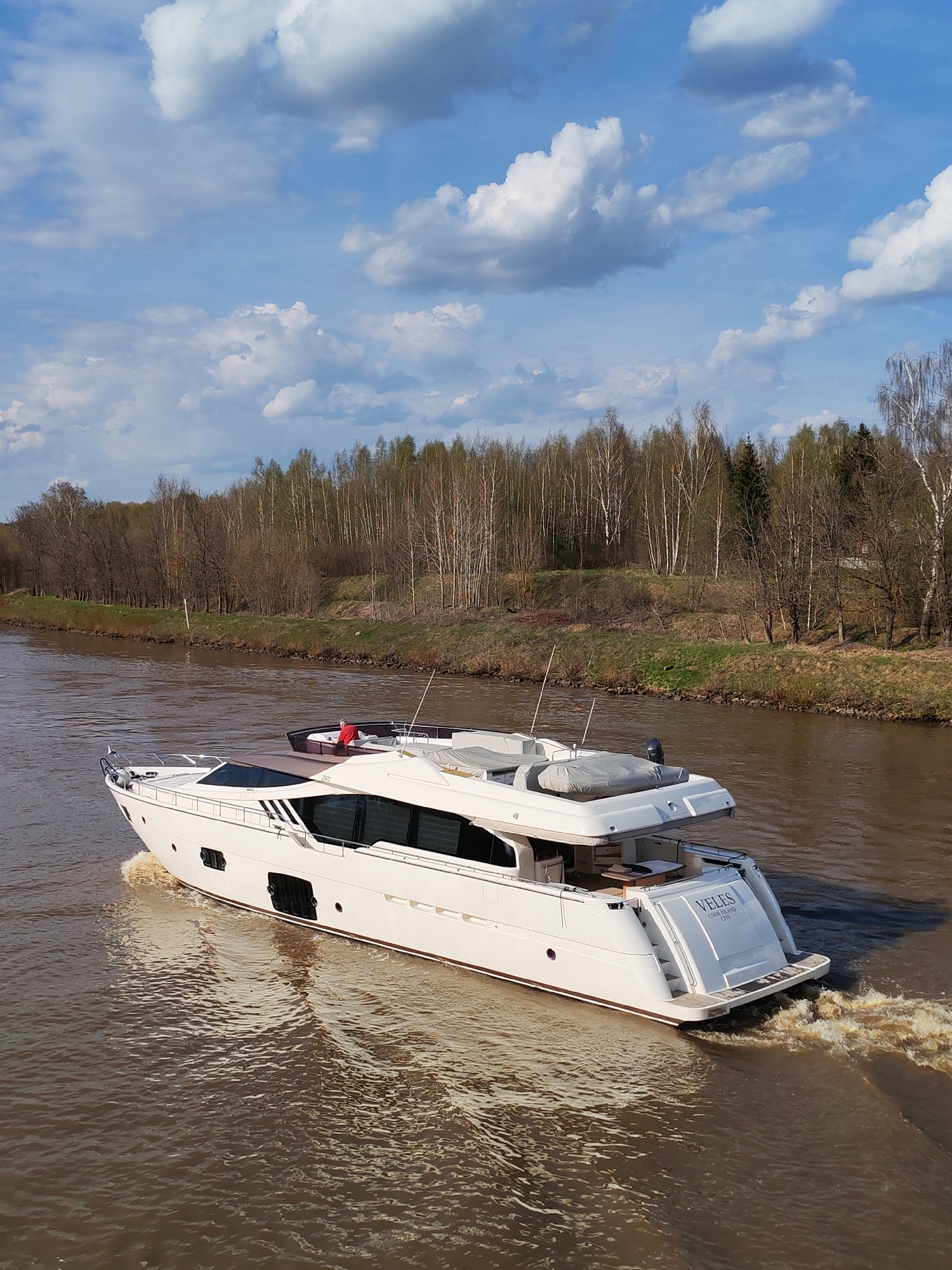 A white motor yacht, named "VELES COOK ISLAND CITS", is underway on a canal in Yakhroma, Russia. The canal water is brown. The banks of the canal have vegetation and bare trees, indicating early spring. A person in a red shirt is visible on the top deck of the yacht. The sky above is blue with scattered clouds.