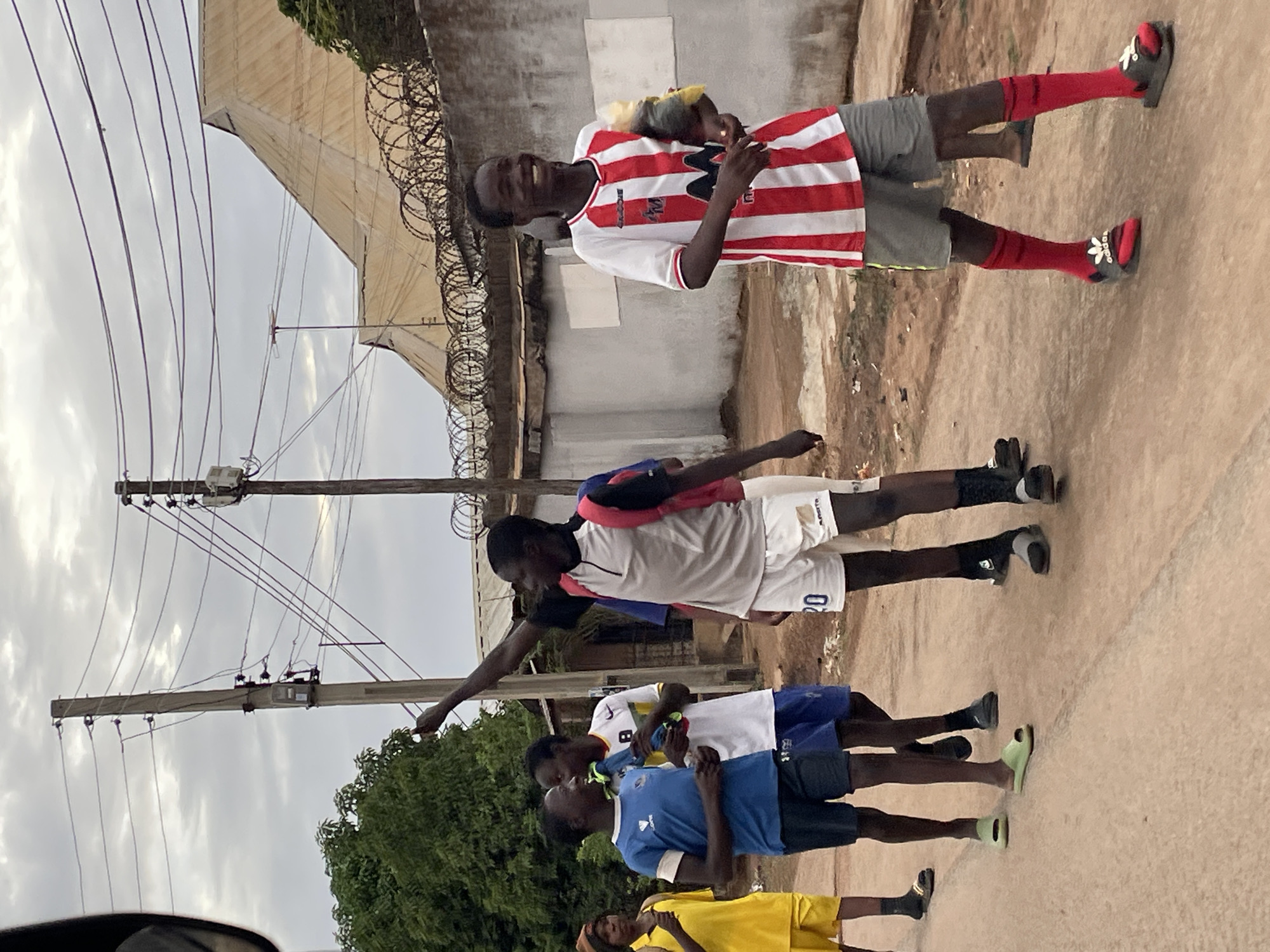 The image shows a group of five young people standing on a paved ground in an urban area. The location is indicated as the city of Jos, Nigeria. One person wears a red and white striped shirt and grey shorts, with red socks and black slides. The second is in a white shirt and white shorts with the number "20" printed on the right leg, wearing black socks and black slides. The next three wear blue, white, and yellow shirts with matching shorts respectively.
The background includes a barbed wire fence atop a concrete wall, a building, electrical poles, and wires against a cloudy sky. Some greenery is also visible. One person has their arm reaching towards the electrical poles.