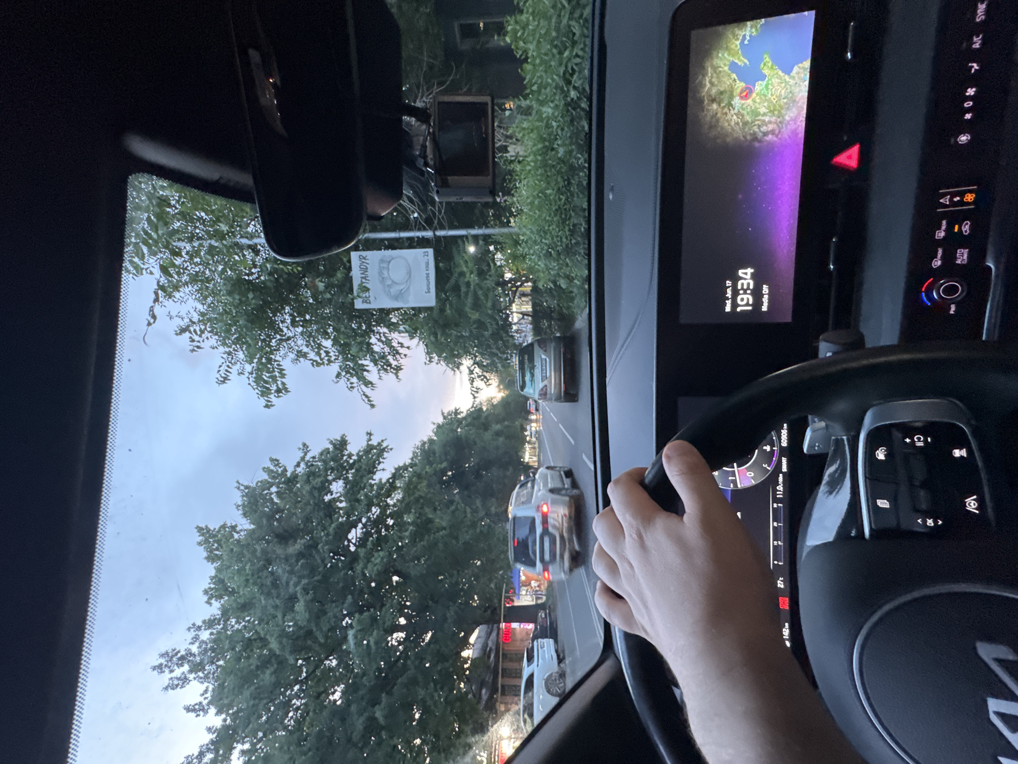 The image shows a first-person view from the driver's seat of a car in Almaty, Kazakhstan. The driver's hand is on the steering wheel, which has a modern digital dashboard display. The dashboard shows the date "Wed, Jun 17", the time "19:34", and a map of a coastal region, possibly the Caspian Sea, with a red marker on a city labeled "Baku". The car is on a street lined with trees, and other vehicles are visible on the road, including a white SUV in front and a white sedan to the left. There are buildings and shops along the street, and a sign with the text "BESTANDYR" is visible hanging from a pole. The sky appears overcast and it is likely late afternoon or early evening.