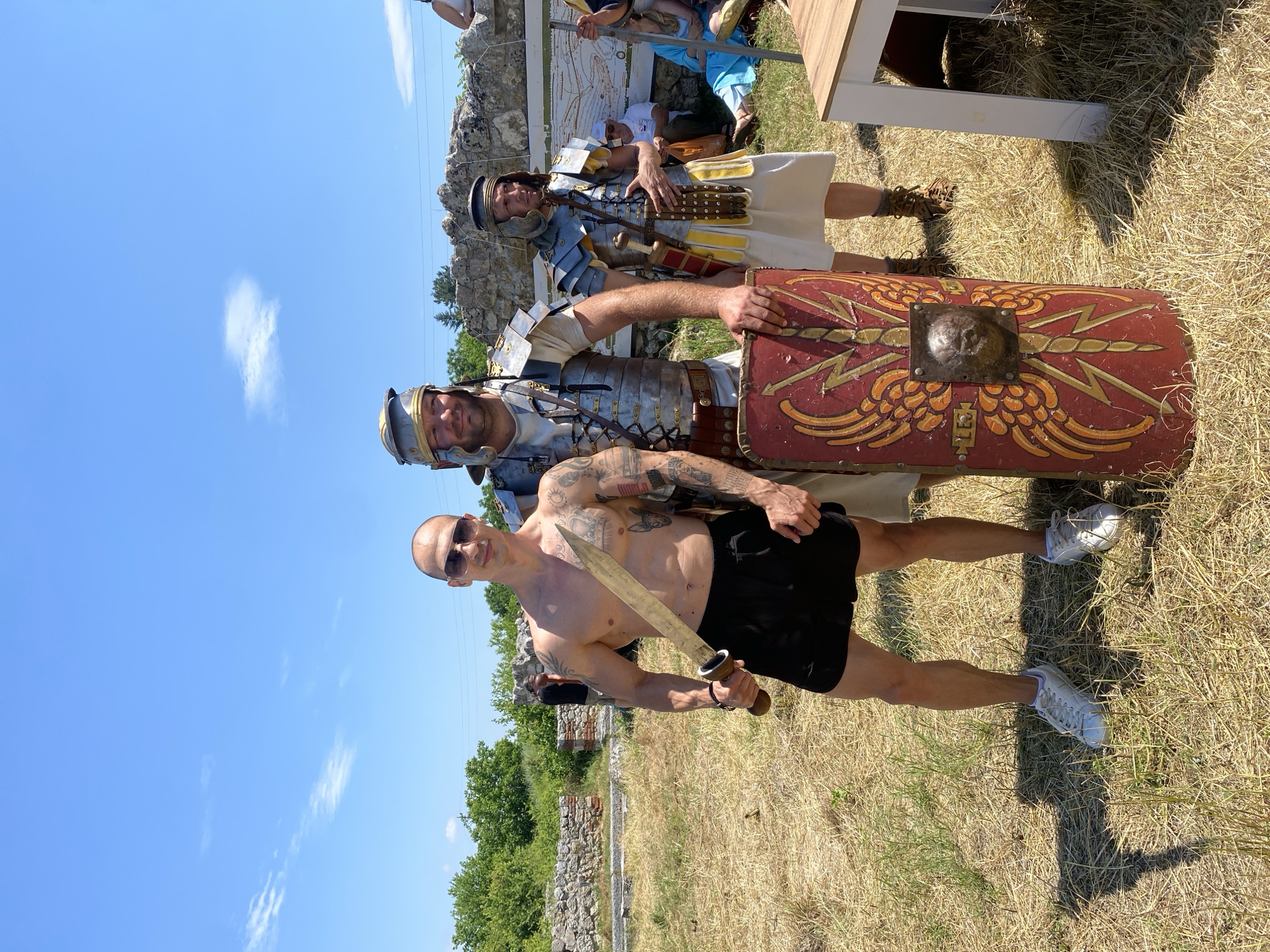 The image depicts three men posing outdoors on a sunny day in Gigen, Bulgaria, against a backdrop of ancient stone ruins and dry, sun-parched grass. The sky is clear blue with a few scattered white clouds.
From left to right, the first man stands shirtless, revealing a muscular physique and numerous tattoos covering his arms and torso. He has a bald head, wears sunglasses, black shorts, and white athletic shoes. He holds a short, bronze-colored sword with a wooden hilt in his right hand, angled slightly upward. His gaze is directed towards the camera.
The second man, positioned centrally, is dressed as a Roman legionary. He wears a silver-colored lorica segmentata (segmented plate armor) over a white tunic, and a helmet. He holds a large, red rectangular scutum (shield) upright, resting on the ground to his left. The shield is decorated with yellow winged lightning bolts and features a central bronze boss embossed with a lion's head. He has a beard and looks towards the camera.
The third man, standing slightly behind and to the right of the second, is also in Roman military attire. He wears a helmet, armor resembling scale or mail over a white tunic, and a pteruges (skirt-like apron) with alternating yellow and white strips. A gladius (short sword) in a scabbard is visible at his left hip. He wears brown leather boots and looks slightly to his left.
In the background, remnants of stone walls and structures are visible, consistent with ancient ruins. Green trees and bushes line the distant horizon. Several other individuals are present in the far right background, partially obscured, including one person in a white hat and another draped in blue fabric. A white table with a wooden top is partially visible in the extreme lower right foreground. Strong sunlight casts distinct shadows on the dry grass.