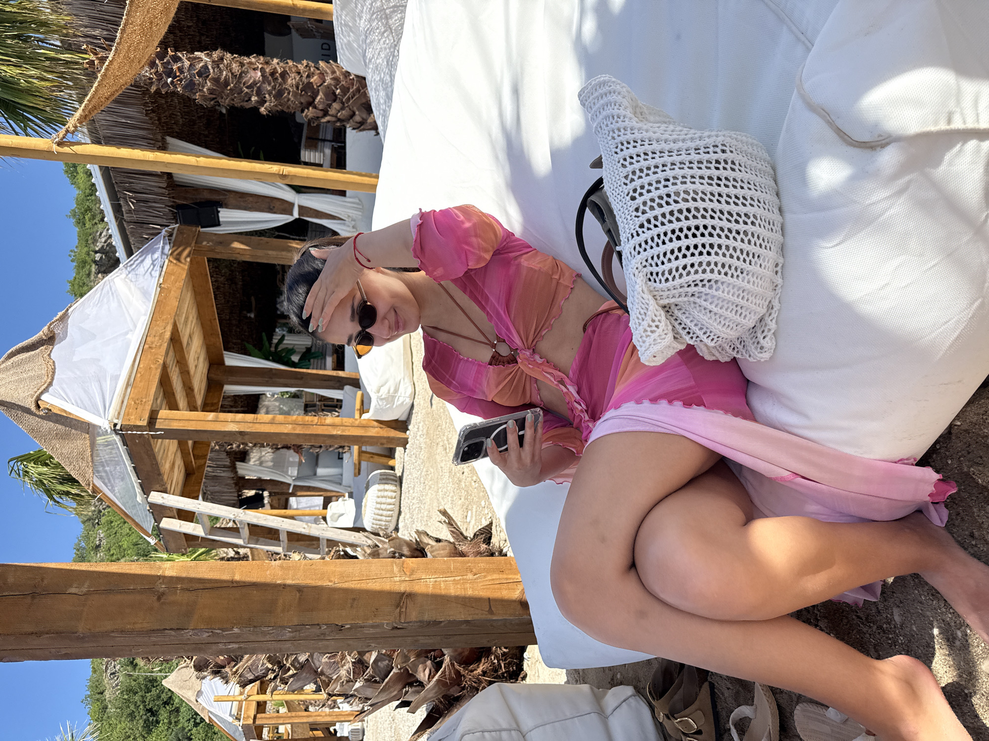 An outdoor scene in Drobnići, Montenegro, depicts a young adult female reclining on a white lounge bed under bright, clear daylight. The subject, dressed in a pink and orange ombre long-sleeved crop top and matching skirt, has dark hair pulled back and wears dark oval sunglasses. She holds a smartphone in her right hand, gazing at its screen, while her left hand is raised to her forehead, potentially shielding her eyes from the sun. A red string bracelet is visible on her left wrist, and her fingernails are painted light green. Her legs are crossed, and her bare feet rest on the sandy ground next to the lounge bed.
Adjacent to the female subject on the lounge bed is a white, open-weave knitted tote bag. On the ground near her feet are three pairs of sandals: a brown pair with gold buckles, a white pair of flat sandals or flip-flops, and a pair of light-colored espadrille-style wedge sandals.
The background features several wooden structures resembling beach cabanas or huts. These structures have robust wooden posts and beams, with roofs constructed from a combination of thatched material and white fabric or clear plastic sheeting. White sheer curtains are draped from some of the structures, and white beanbag-style lounge cushions are visible within and around them. A wooden ladder leans against one of the structures on the left. Palm trees with textured dark brown trunks are interspersed throughout the setting. In the distance, lush green hills rise under a clear blue sky. The ground surface is sandy or gravely.