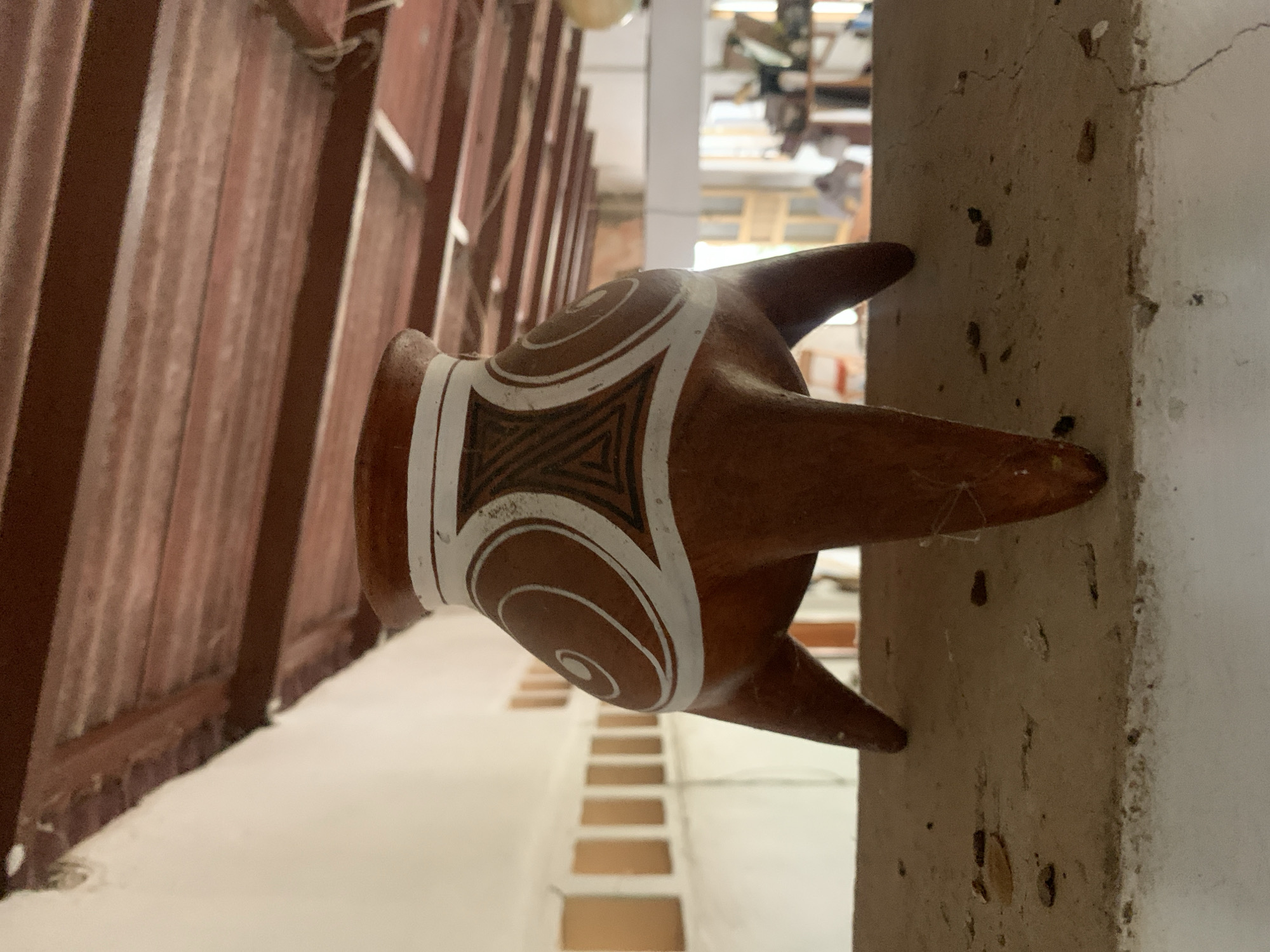 This image, captured in Santa Ana, Venezuela, showcases a decorative, brown clay tripod vessel. The pottery piece is adorned with distinct white geometric patterns, featuring swirling spirals on its rounded body and a central rectangular band filled with black and white triangular motifs. Three robust legs extend from the base of the vessel, giving it a unique silhouette.
The vessel is situated indoors or within a sheltered area. To its left, a reddish-brown wooden structure, possibly a wall or partition, is visible, constructed from vertical planks. To its right, a sturdy, light-colored concrete or stucco column displays surface cracks and minor dark spots, suggesting age or natural wear. The background is softly blurred, revealing architectural elements such as a series of light-colored steps or tiers receding into the distance. The lighting is diffused, suggesting a daytime setting. No individuals or specific activities are depicted in the scene, which primarily focuses on the static arrangement of the pottery within its environment. There is no visible text.