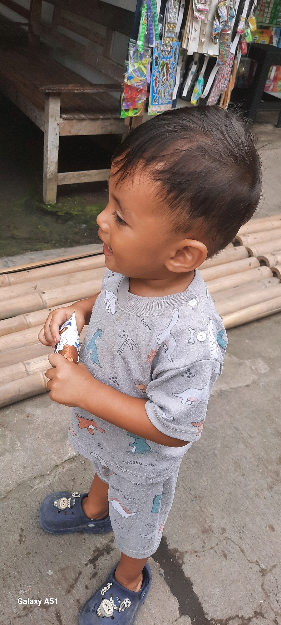 A young child, likely a toddler, stands outdoors holding a wrapped ice cream bar. The child is wearing a gray t-shirt and shorts set with a dinosaur pattern, and dark blue clogs with character embellishments. The child's hair is dark and short, and they are looking to the left of the frame with their mouth slightly open. The child is standing on a concrete surface, with a row of bamboo poles visible behind them. In the background, a wooden bench is partially visible, and a display of various items, possibly toys or snacks, hangs on a rack. The scene appears to be in a casual outdoor setting, potentially a market or a roadside stall, given the merchandise display. The presence of a "Galaxy A51" watermark in the bottom left corner suggests the image was captured with a Samsung Galaxy A51 smartphone. The location is identified as Ploso, Indonesia.