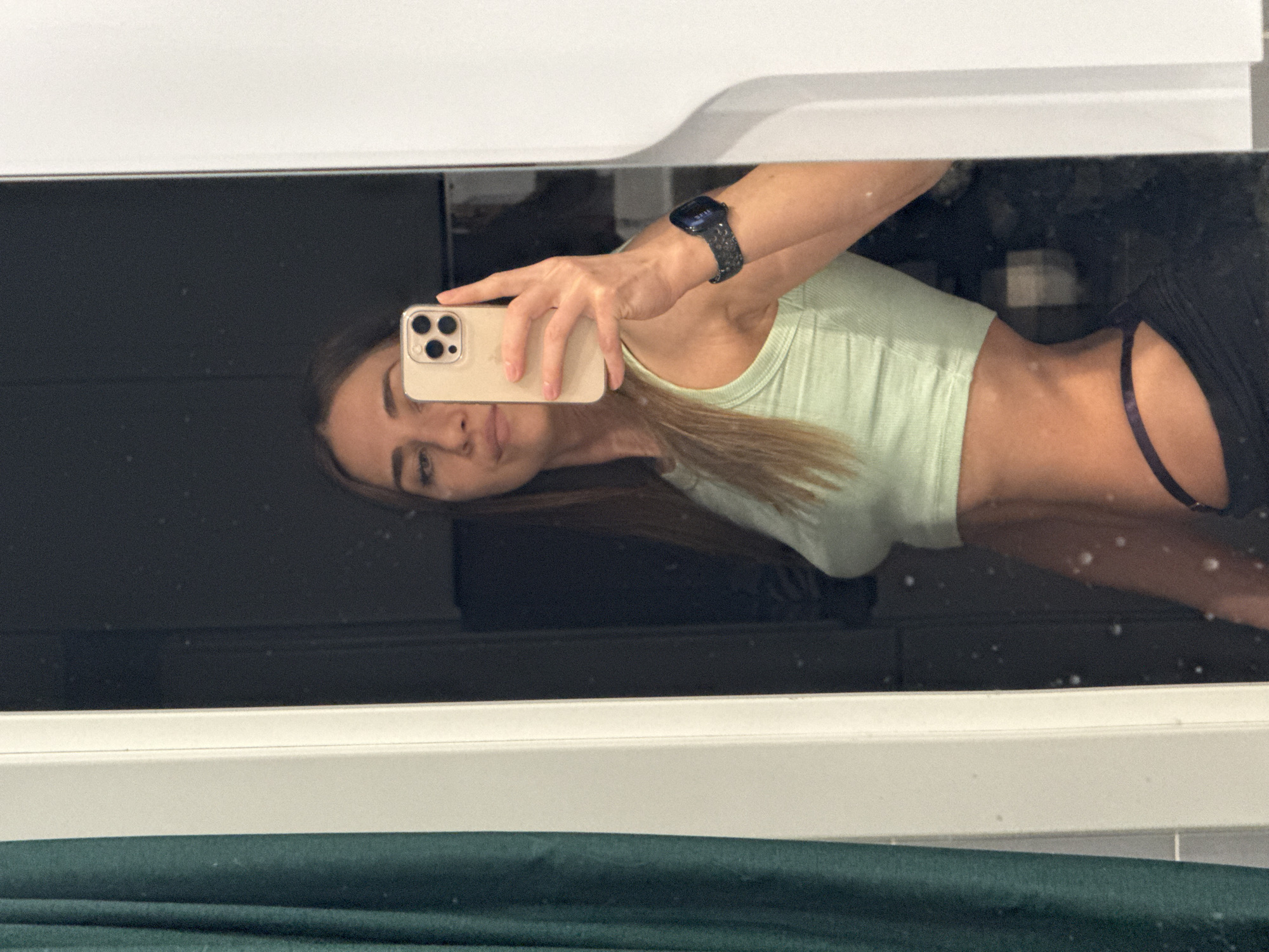 A mirror selfie captures a woman wearing a light green tank top and dark shorts. She is holding a smartphone in her hand, and a watch is visible on her wrist. The reflection shows a bathroom setting, with a white frame surrounding the mirror. The bottom of the frame overlaps a greenish-blue surface, possibly a countertop or fabric. There's no information available to determine whether the photo was taken in Lyubertsy, Russia.