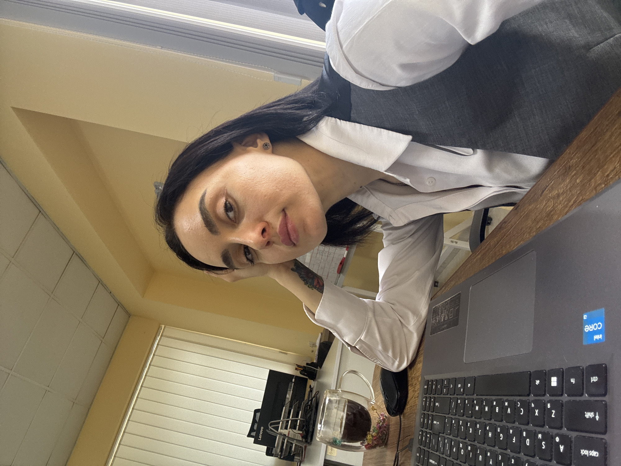 A woman with long black hair is seated at a wooden desk with a laptop. She is wearing a white blouse and a grey vest. Her left arm is resting on the desk, revealing a tattoo on her forearm. A glass mug filled with a dark liquid and small colorful flowers sits to the left of the laptop. In the background, a computer monitor and office equipment are visible. The setting appears to be an office space with a window, possibly located in Tbilisi, Georgia. The picture is taken at an angle, as the photographer seems to be positioned slightly above and to the side of the woman.