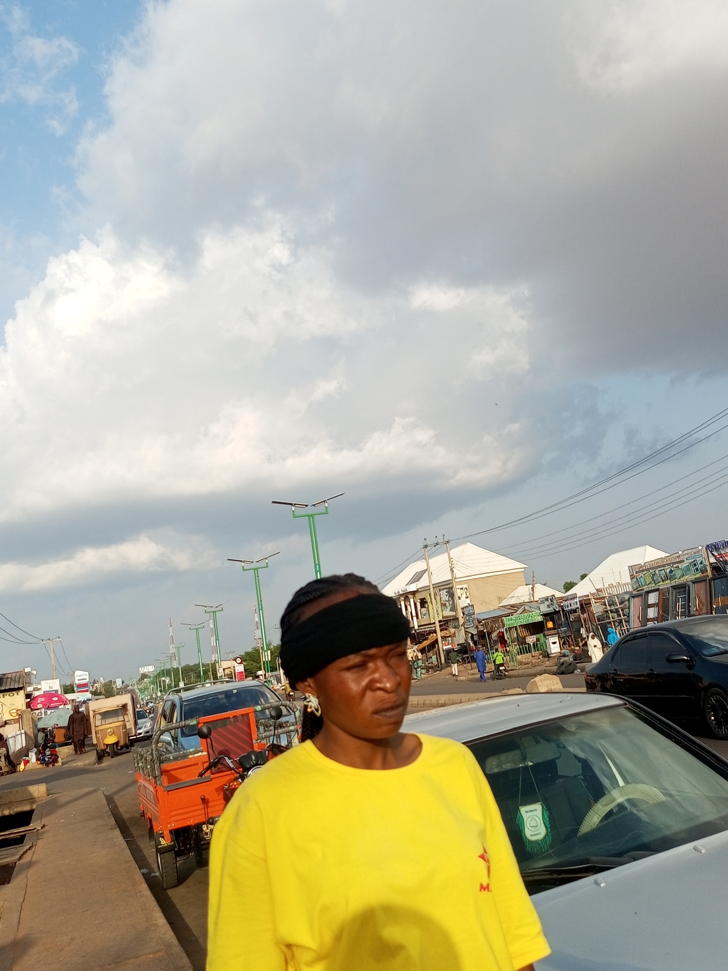 The image captures a street scene in Tempure, Nigeria, likely during the late afternoon or early evening due to the warm lighting and diffused sunlight. The sky is a prominent feature, filled with a mix of bright white and grey clouds, suggesting an overcast or partly cloudy condition.
In the foreground, a woman is partially visible on the right side of the frame. She is wearing a bright yellow t-shirt and a black band across her forehead, obscuring her eyes. Her expression appears somewhat serious or perhaps focused. She is standing next to a silver car, with part of the car's interior, including the steering wheel and a hanging air freshener, visible.
The background depicts a busy street with various vehicles and market stalls. An orange three-wheeled vehicle, often used for transportation or hauling goods in this region, is parked on the left side of the road. Further down the street, several other vehicles, including cars and what appear to be smaller commercial tricycles, are visible. Streetlights with solar panels are lined up along the road, indicating urban infrastructure. Buildings with light-colored roofs line the street, and market stalls with various goods are set up along the roadside. People are seen walking and interacting, suggesting daily commercial activity. One of the market stalls has visible text, but it is too small and indistinct to be read. Another sign on a building on the left appears to have some lettering as well. The overall scene conveys a sense of a bustling local marketplace and transportation hub in Nigeria.