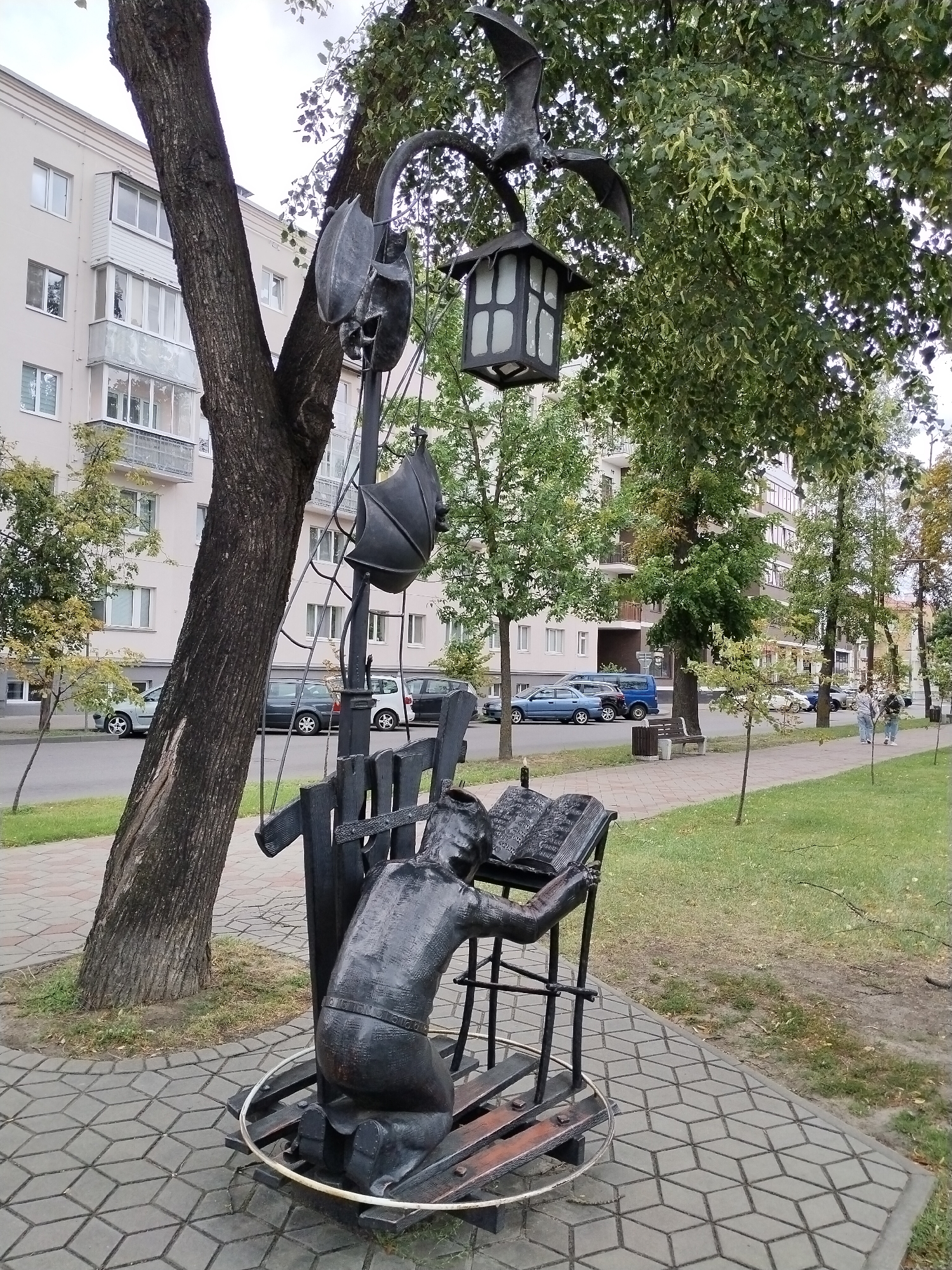 The image depicts a street scene in Brest, Belarus, featuring a public art installation. The primary subject is a dark-colored metal sculpture of a person kneeling before an open book. The sculpture is positioned on a circular base with wooden planks, and a metal fence-like structure surrounds it. Above the sculpture, a lamppost is visible, adorned with decorative elements that resemble bats and a lantern. The lamppost is situated next to a large tree with textured bark.
In the background, a multi-story apartment building with balconies and windows lines the street. Cars are parked along the road, and a sidewalk paved with hexagonal tiles runs alongside a grassy area. Several trees with green foliage are planted along the sidewalk. Two individuals, a woman and a man, are walking on the sidewalk in the distance, facing away from the camera. The overall environment suggests an urban park or a well-maintained street in a residential area. The sky appears overcast.