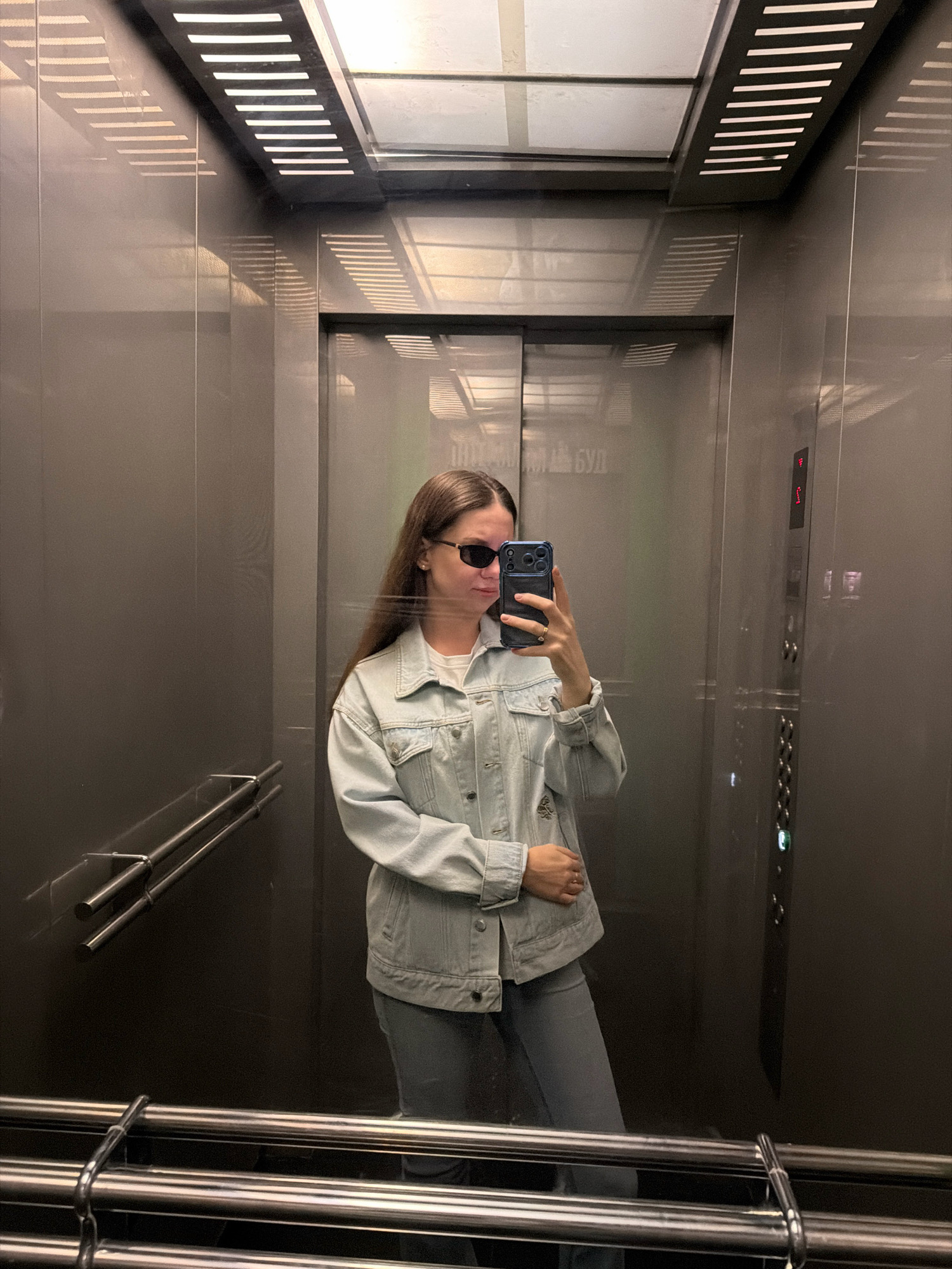 A person with long brown hair, wearing sunglasses, a light blue denim jacket, and grey pants, stands in an elevator and takes a selfie with a smartphone. The elevator has grey metallic walls and is lit by overhead fluorescent lights, casting linear shadows on the walls. A set of metal handrails is visible on the left wall, and a control panel with buttons is on the right wall. The background reflection suggests an urban environment, potentially the city of Kyiv, Ukraine, given the visible Cyrillic text on the elevator doors.