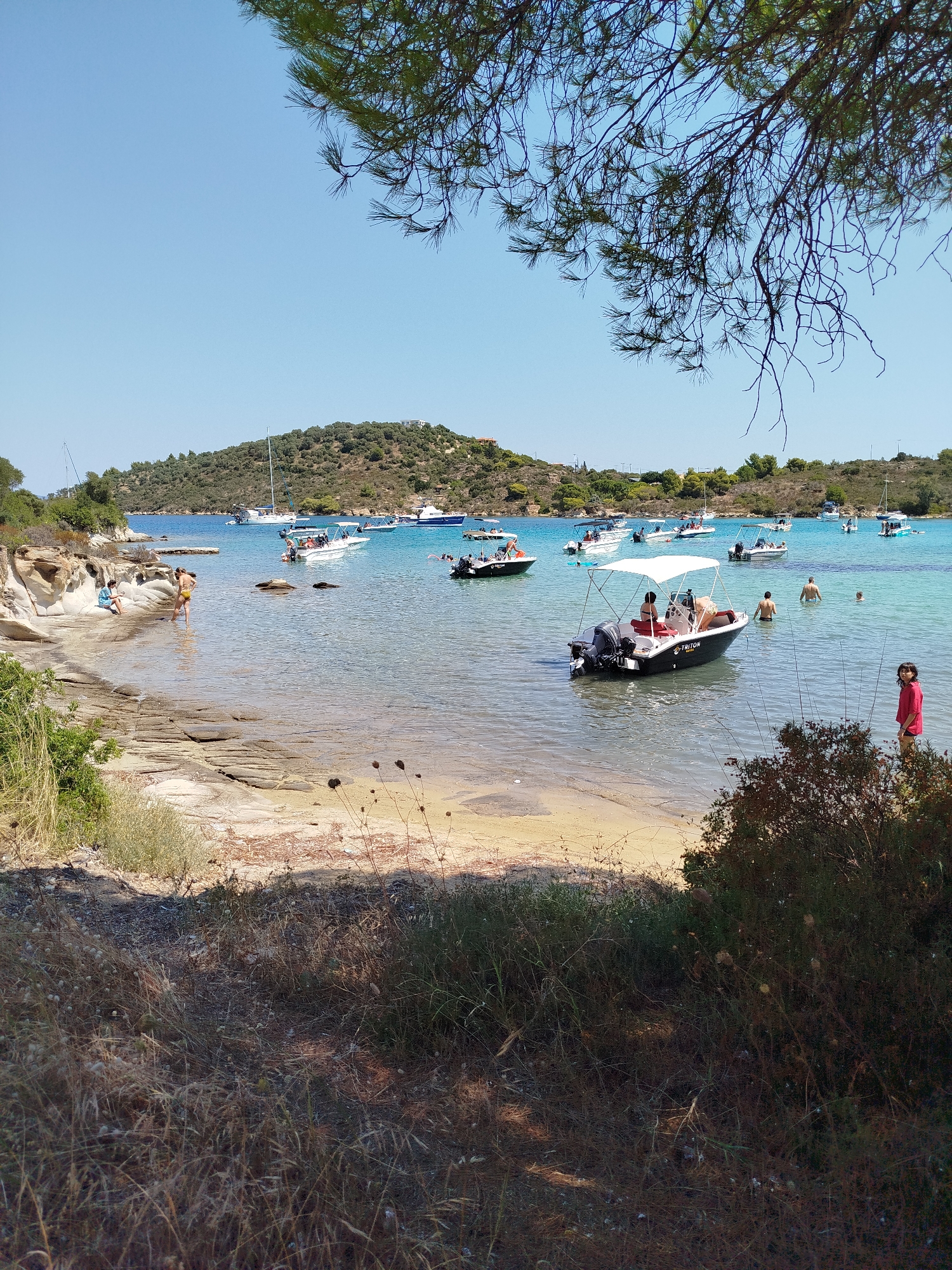 The image depicts a sunny day at a beach in the Sithonia Municipal Unit, Greece. The scene is characterized by clear turquoise waters, a sandy and rocky shoreline, and lush green hills in the background. Several boats, including speedboats and sailboats, are anchored in the bay, with some people swimming and wading in the shallow water. On the shore, two individuals are sitting on rocks, while others are standing or walking near the water's edge. Vegetation, including dry grasses and a dense bush, frames the foreground of the image. The sky is a clear, bright blue.