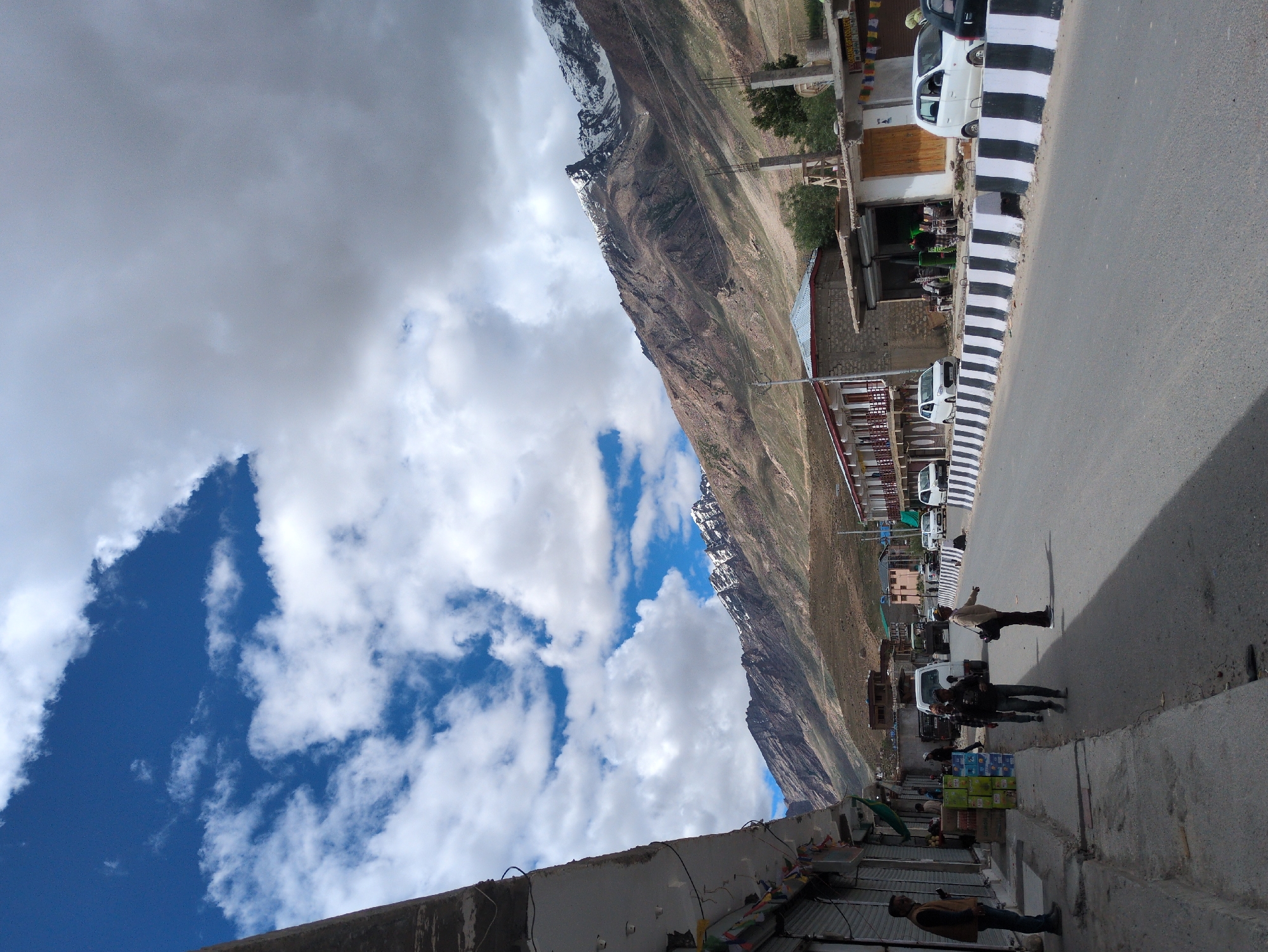The image depicts a street scene in the town of Padum, India, with a backdrop of majestic, snow-capped mountains. A paved road runs through the center of the frame, bordered by a concrete barrier painted with black and white stripes. On the left side of the road, a row of shops with closed shutters lines the street, with prayer flags strung across the front of one establishment. Several people are visible on the sidewalk and street, including a man standing near a shop, two individuals walking down the sidewalk, and another person in the street interacting with the scene. Several cars are parked along the road and in front of the shops. In the background, the rugged, brown slopes of the mountains rise dramatically, with patches of snow glistening on their peaks under a dynamic sky filled with white clouds against a vibrant blue.