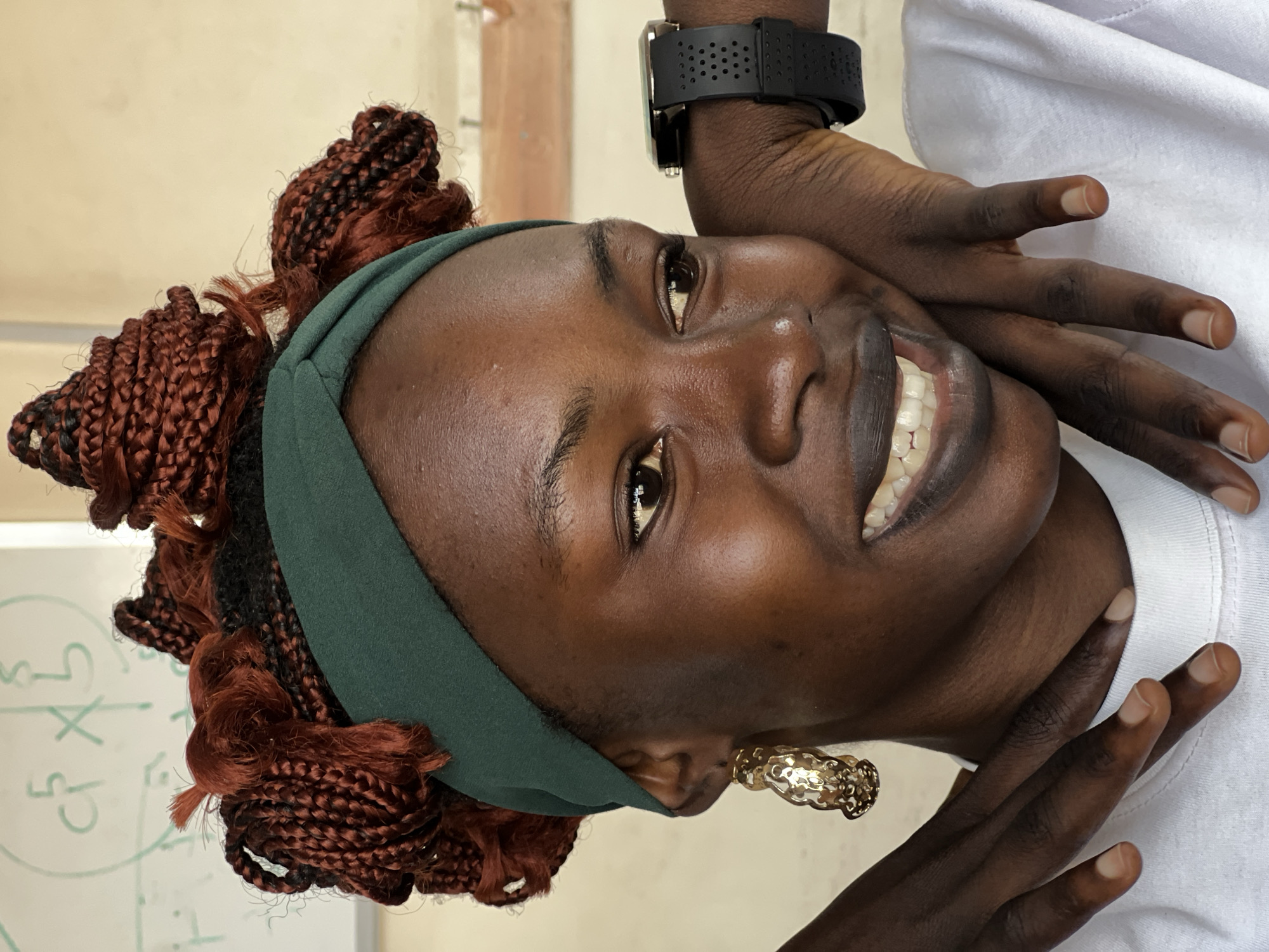 A medium shot shows a young Black woman with dark skin and braided auburn hair, pulled up and styled in a bun, wearing a dark green headband. She has a wide smile, revealing white teeth, and is looking off to the right of the frame. Her left hand is resting on her chin, and her right arm is resting on her shoulder. She is wearing large, gold, ornate earrings and a black smartwatch. She is wearing a white t-shirt. In the background, a whiteboard with some mathematical equations is visible. The lighting is natural and soft, suggesting an indoor environment. The image is cropped tightly around the woman, with no clear indication of the specific location within Jos, Nigeria, though the presence of a whiteboard and classroom-like setting hints at an educational institution.