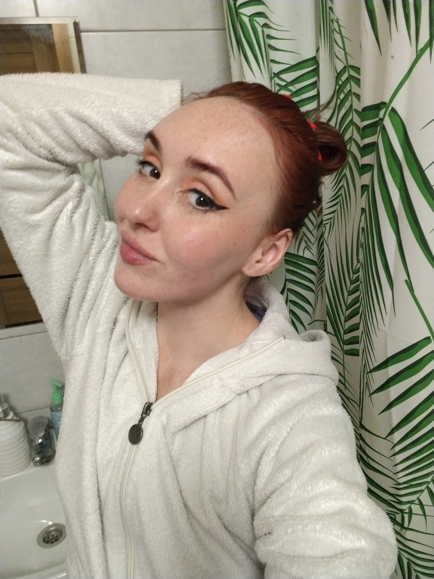 A selfie taken in a bathroom. A woman with red hair, styled in a bun secured with red bands, is wearing a white, hooded, zippered robe. She is posing with her left arm raised behind her head. She has fair skin with freckles and is wearing makeup, including eyeliner. Behind her is a shower curtain with a green palm leaf pattern. To the left, a white sink and bathroom fixtures are visible. The location in Izhevsk, Russia, is not discernible from the image.