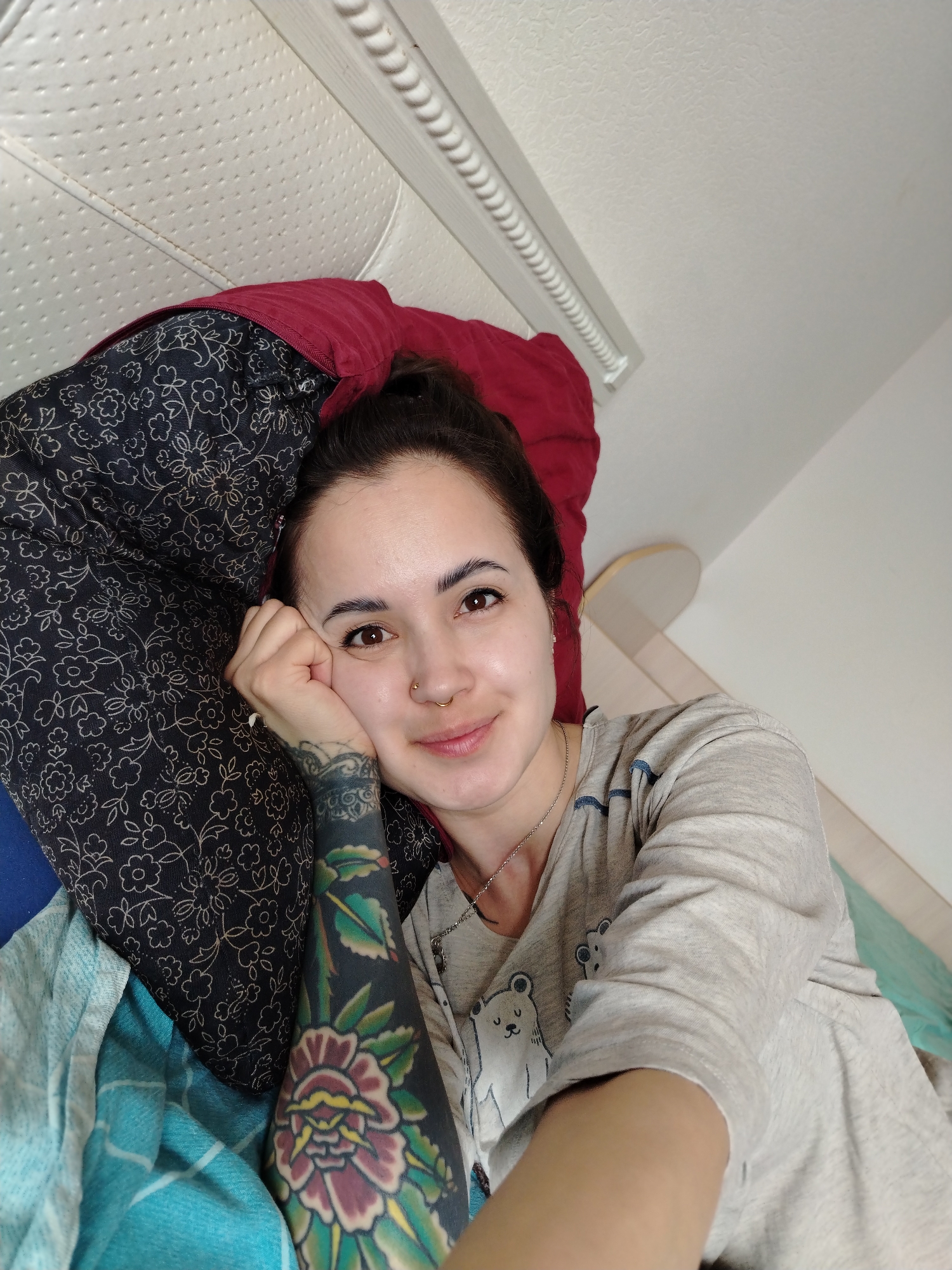 A young woman is pictured in a close-up, slightly high-angle selfie, lying down in what appears to be a bed in Noviy, Russia. She looks directly at the camera with a gentle, relaxed smile. Her dark hair is pulled back, and she has brown eyes. She has a nose piercing, with a gold hoop on her left nostril and a small stud on her right. She wears a light gray t-shirt adorned with cartoon bear graphics and a thin silver chain necklace. Her left arm is heavily tattooed with a colorful full-sleeve design, prominently featuring a large flower with red, yellow, and green elements, surrounded by dark blackwork.
She rests her head on a dark pillow with a light floral pattern, supported by a solid red pillow underneath. A light blue blanket or sheet is visible in the lower left corner of the frame. Behind her head is a white headboard, possibly upholstered with a textured or quilted pattern, topped by a decorative white carved wooden rail. The lighting is soft and even, suggesting an indoor daytime setting. No visible text is present in the image.